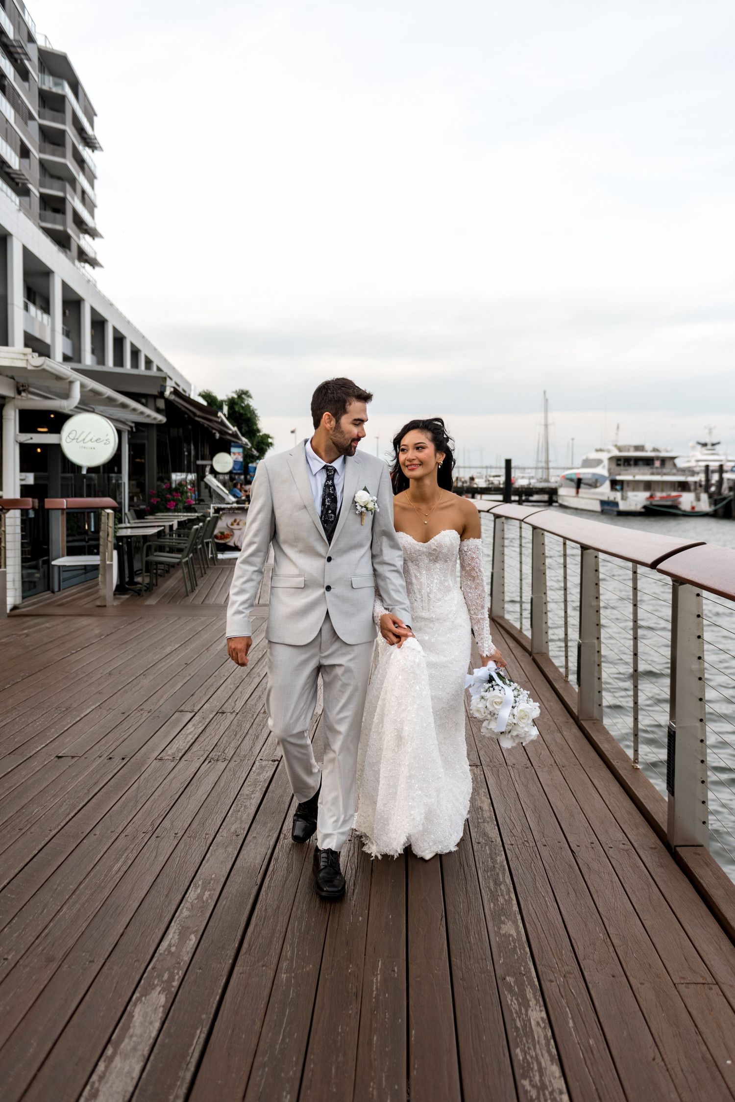 Smiling newlyweds walk hand-in-hand along a waterfront boardwalk with a marina in the background.