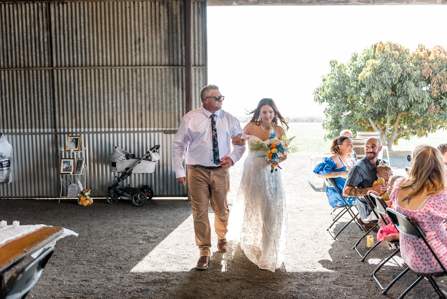 A bride in a lace gown holding a colorful bouquet walks down the barn aisle escorted by a man in a pink shirt.
