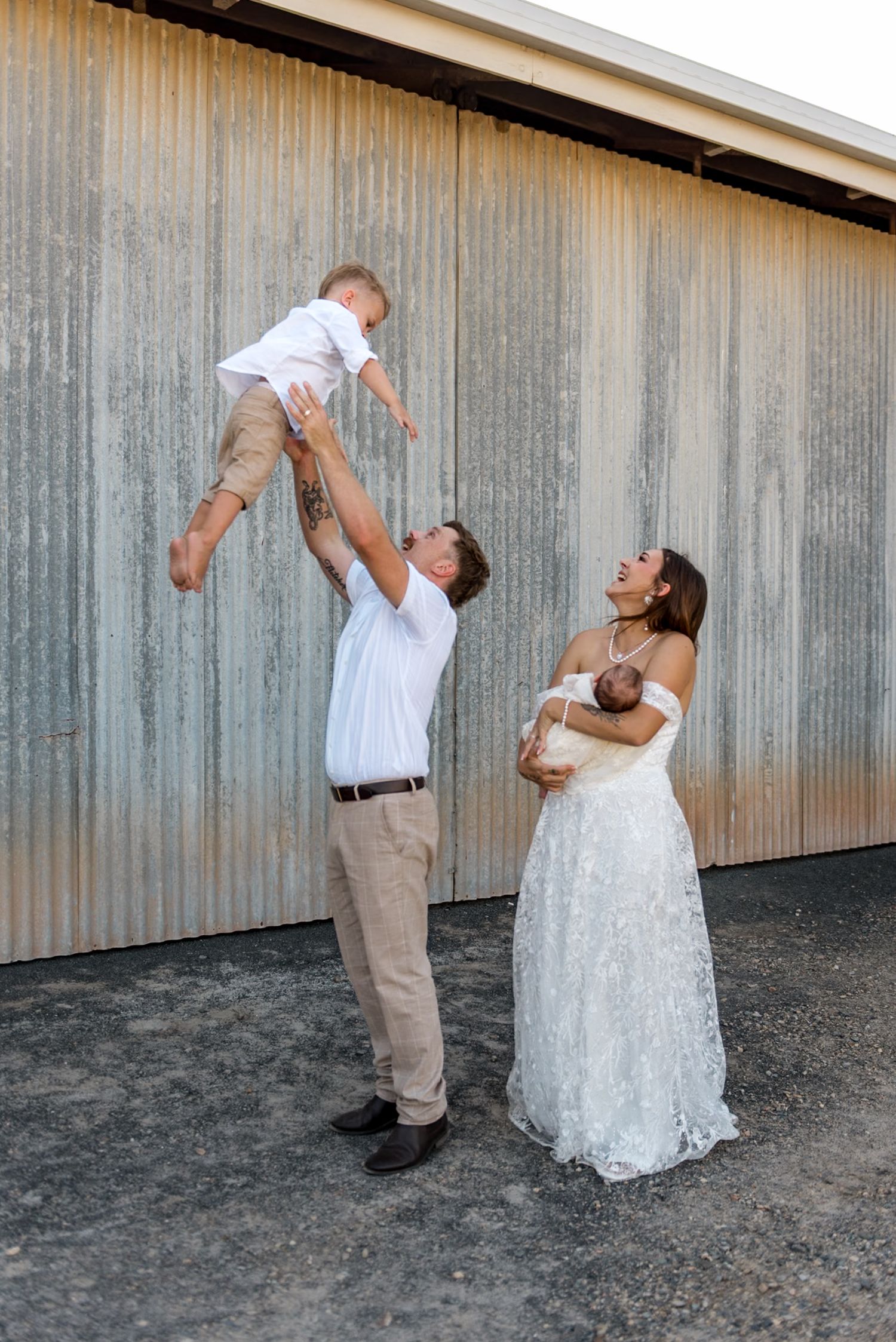 Groom tossing toddler in air while bride in lace wedding dress holds newborn baby, family photo session.