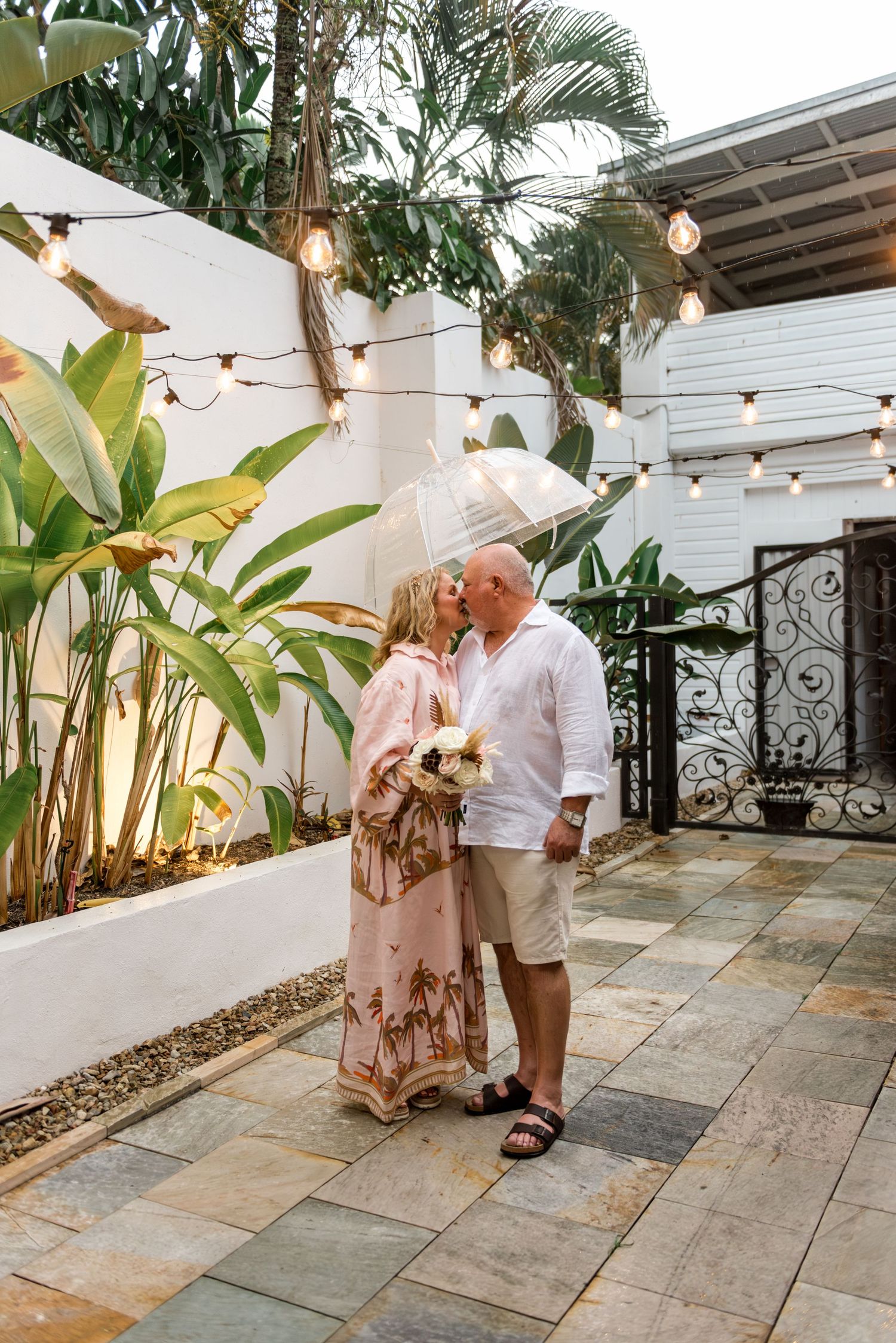 Bride and groom sharing a kiss under clear umbrella in romantic tropical courtyard with festoon lighting.