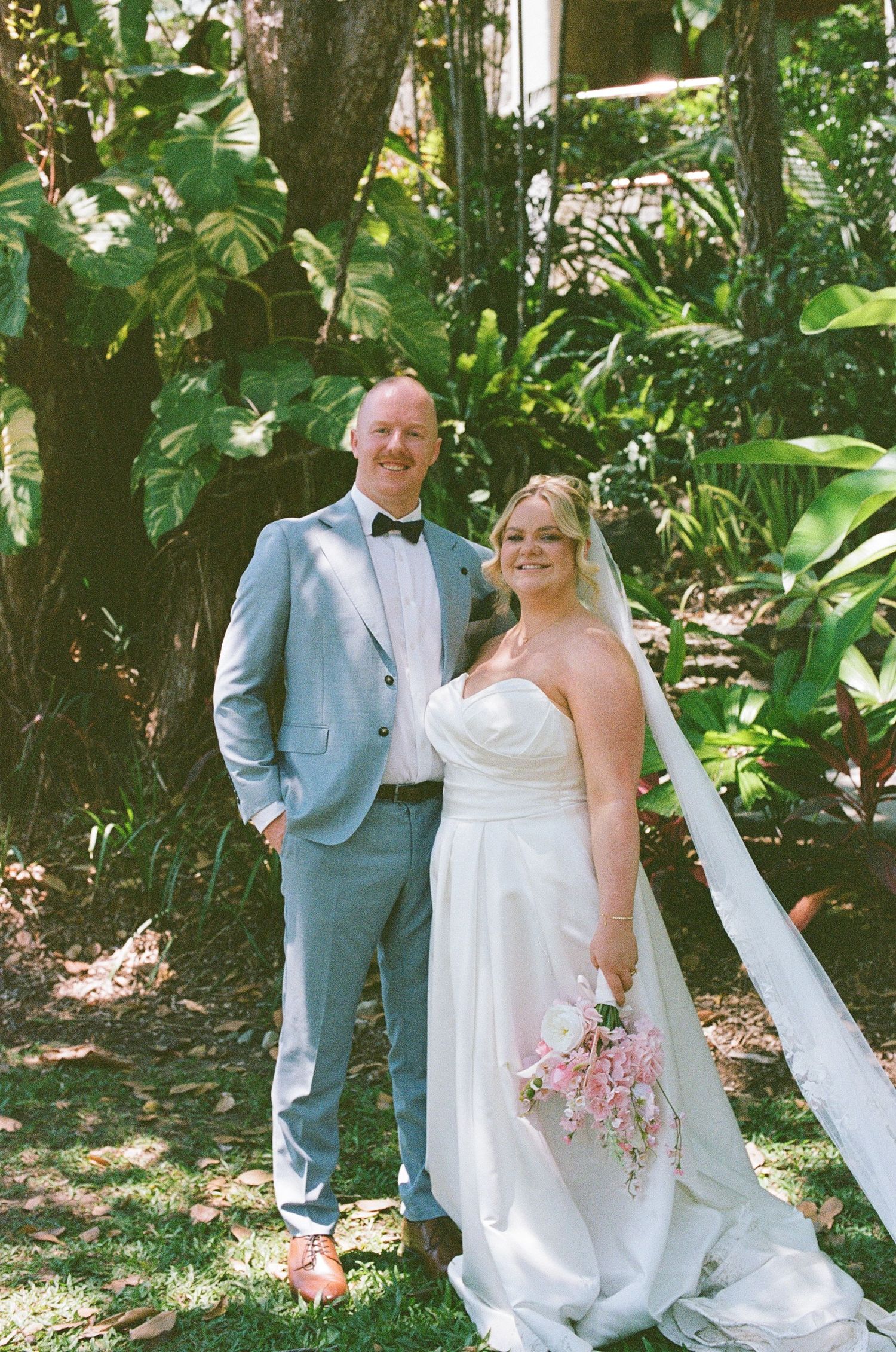 Smiling bride and groom pose together in lush tropical garden, bride holding pink bouquet.