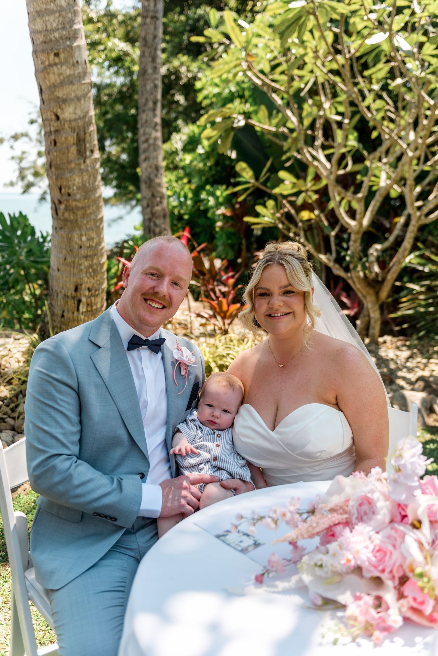 Happy newlywed couple in grey suit and white gown holding infant baby at tropical outdoor wedding ceremony.