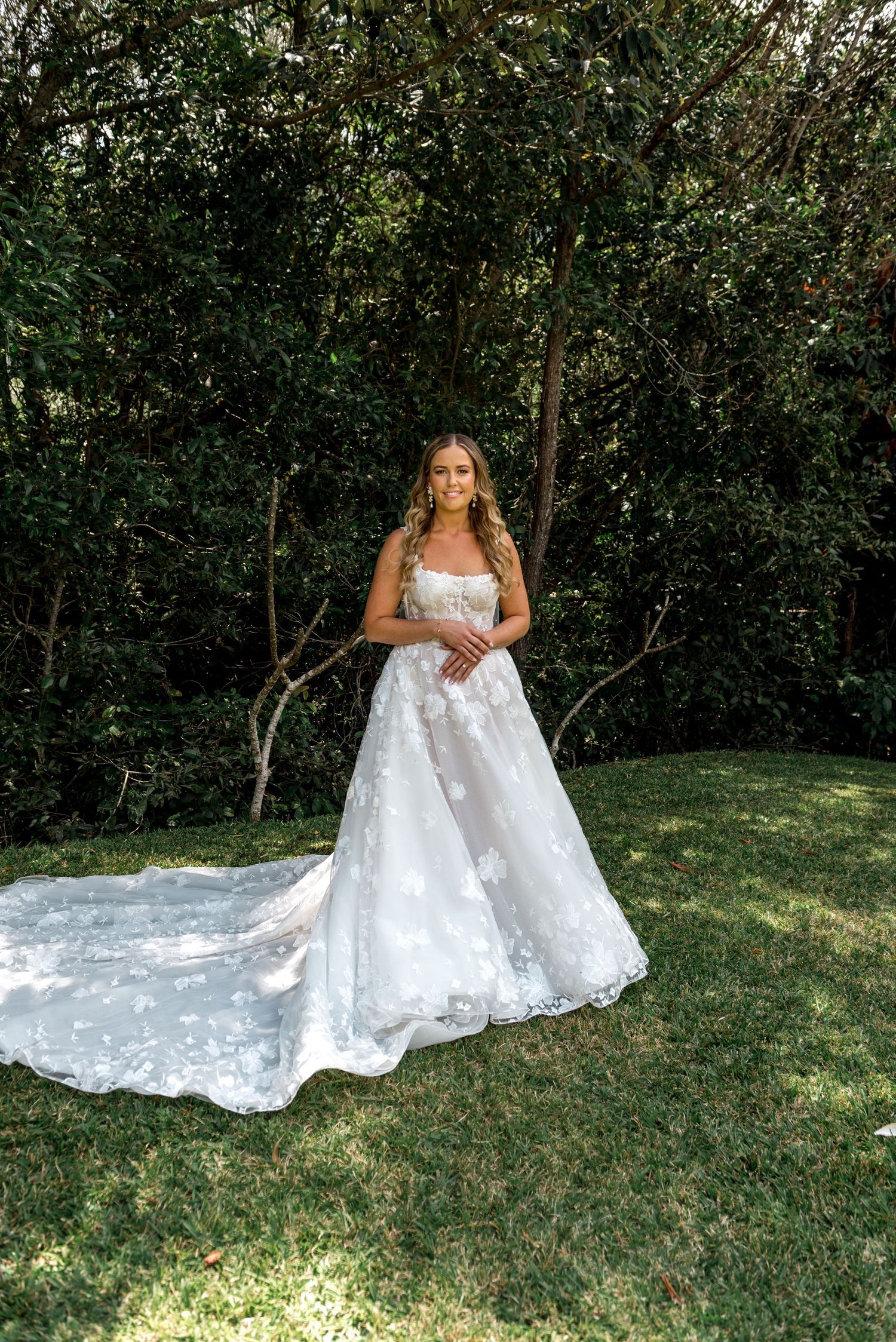 Bride in a floral lace A-line wedding gown with long train standing on green grass before lush trees.