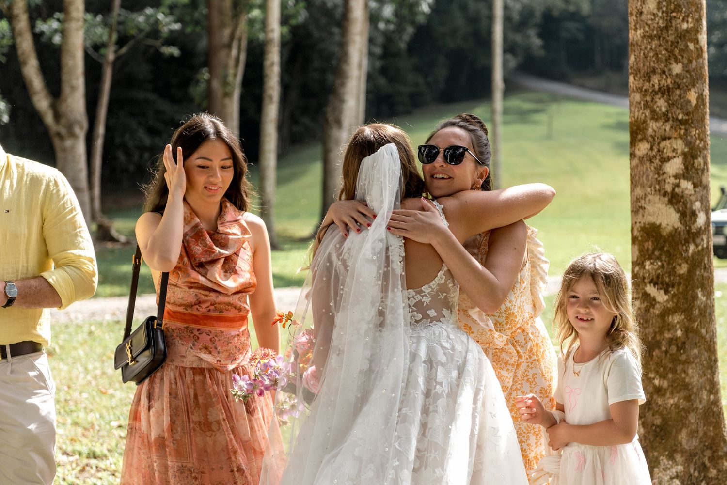 Bride in white gown receiving warm hugs from guests at an outdoor woodland wedding celebration.