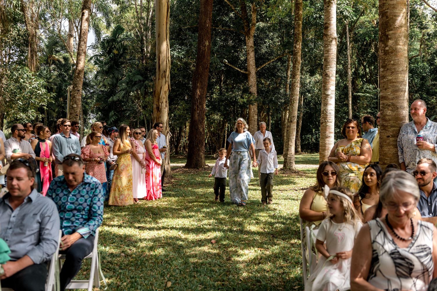 Wedding guests seated in white chairs watch the bridal procession through a lush garden aisle.