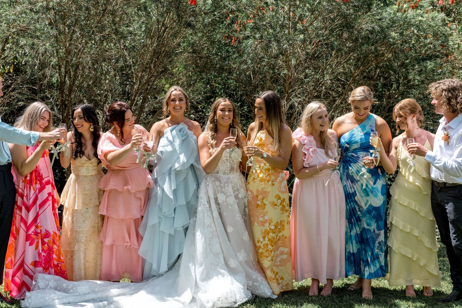 Wedding party in colorful mismatched gowns celebrate with champagne glasses among garden greenery.