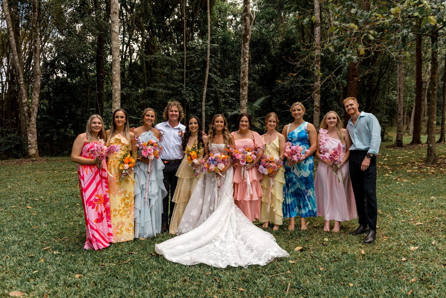 Wedding party posing in colorful mismatched bridesmaid dresses with bright bouquets in forest setting.