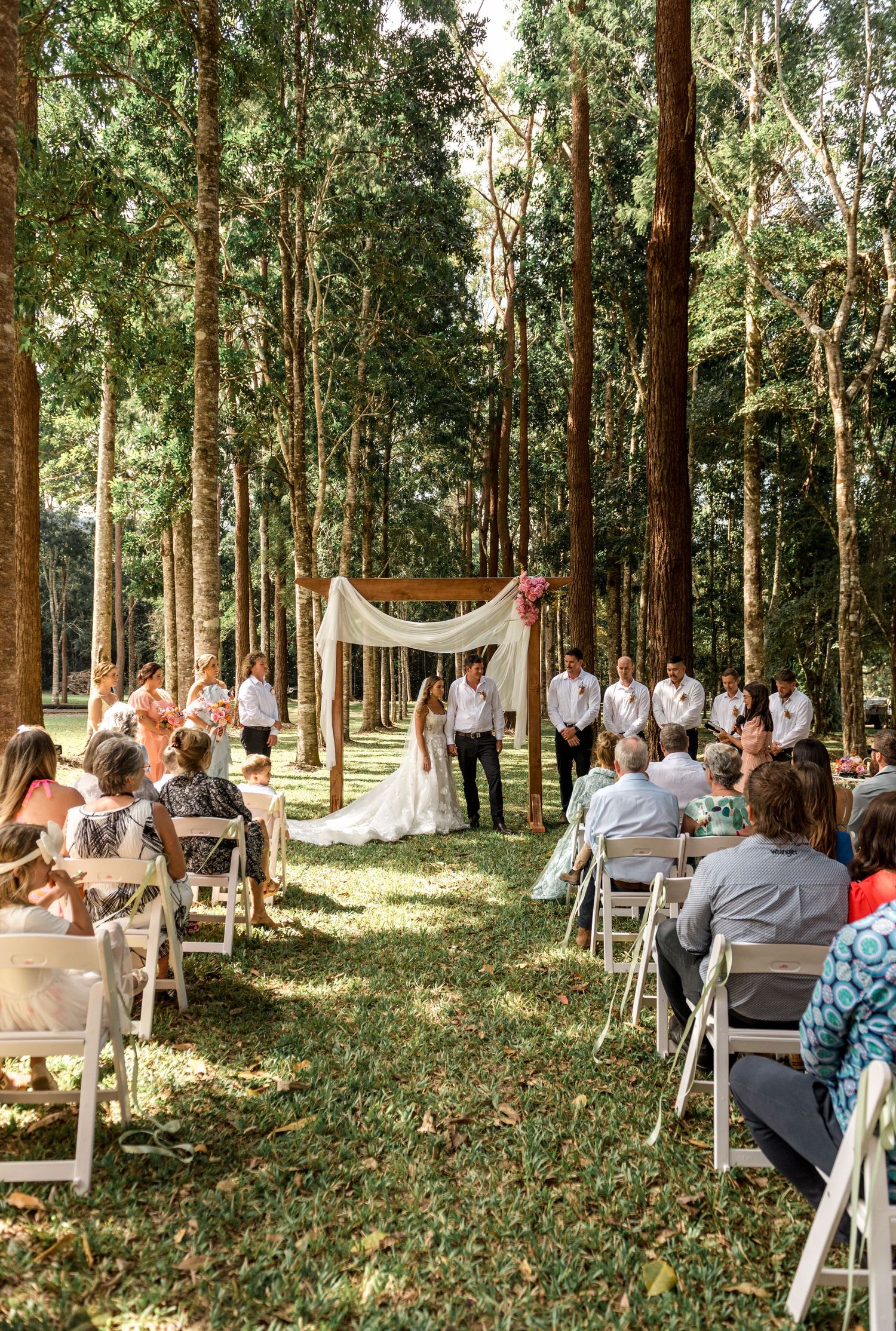 Outdoor wedding ceremony in forest with guests seated on white chairs facing wooden arch altar.