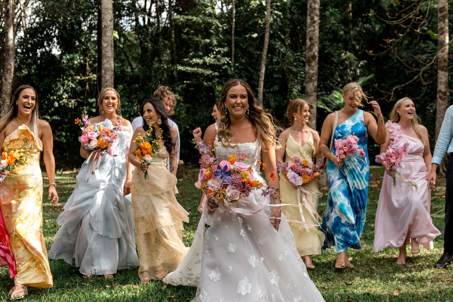 Bride and bridesmaids in vibrant colorful dresses walking joyfully through lush green outdoor venue.