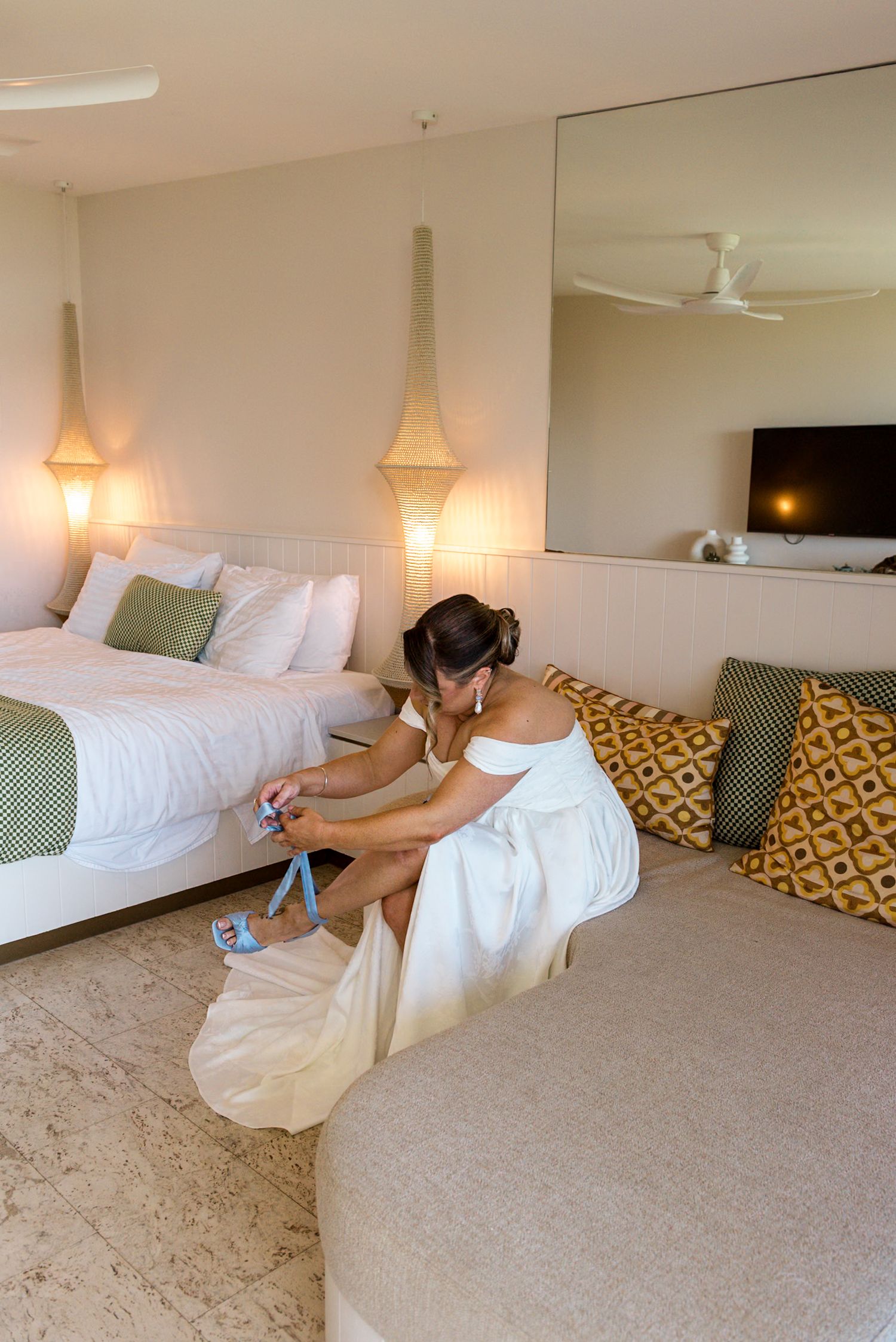Bride in white off-shoulder wedding gown fastening her blue heeled sandals while seated on a hotel couch.