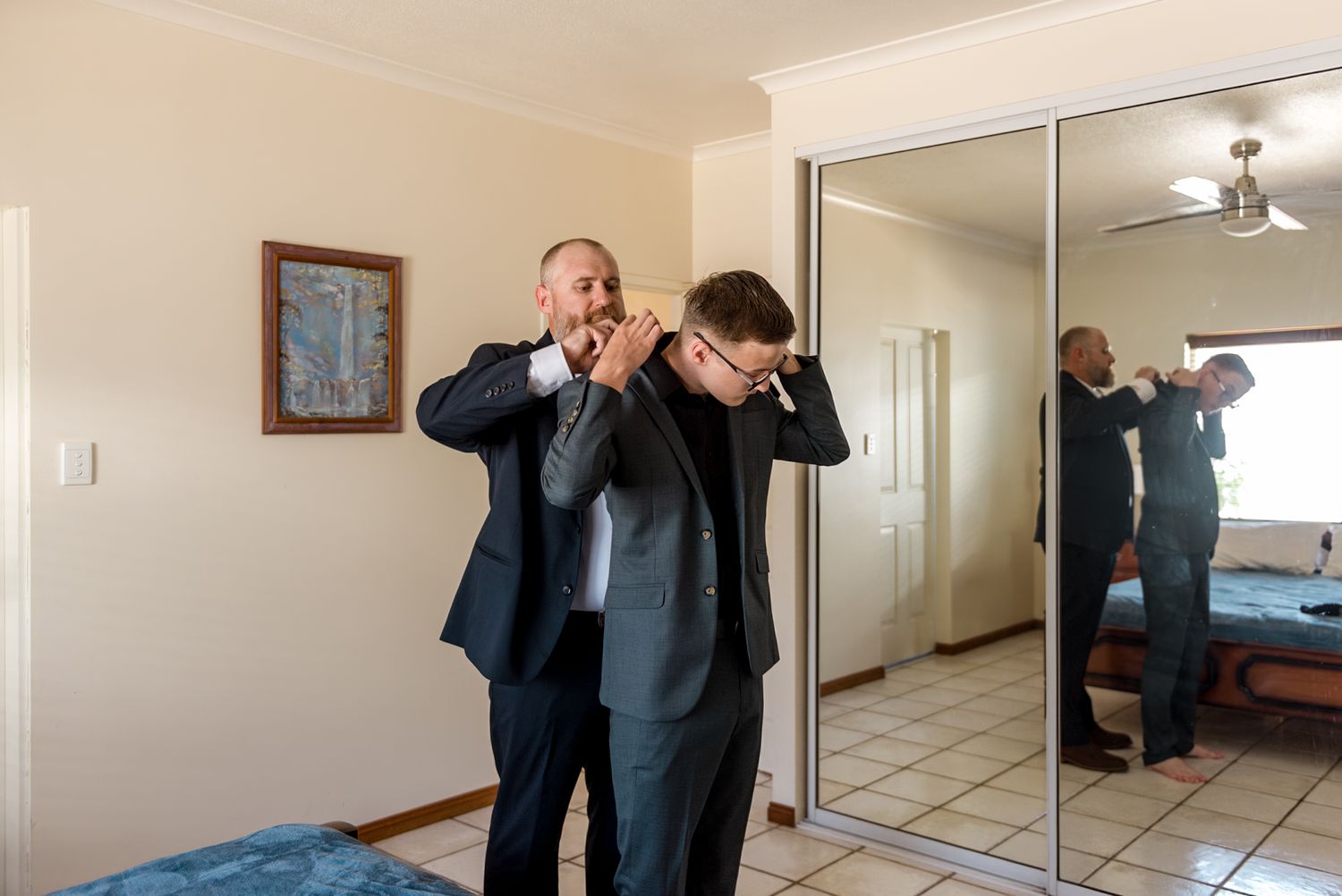 Groom helping groomsman adjust tie in front of mirrored wardrobe while getting ready.