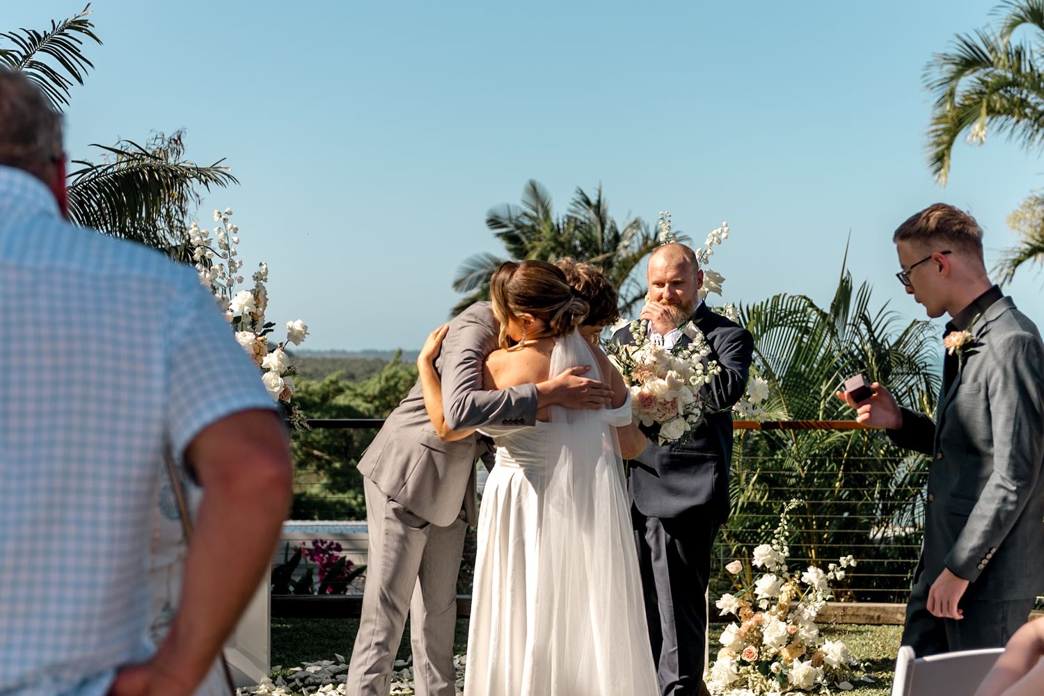 Emotional wedding moment as bride and groom share a hug with family member at outdoor tropical ceremony.