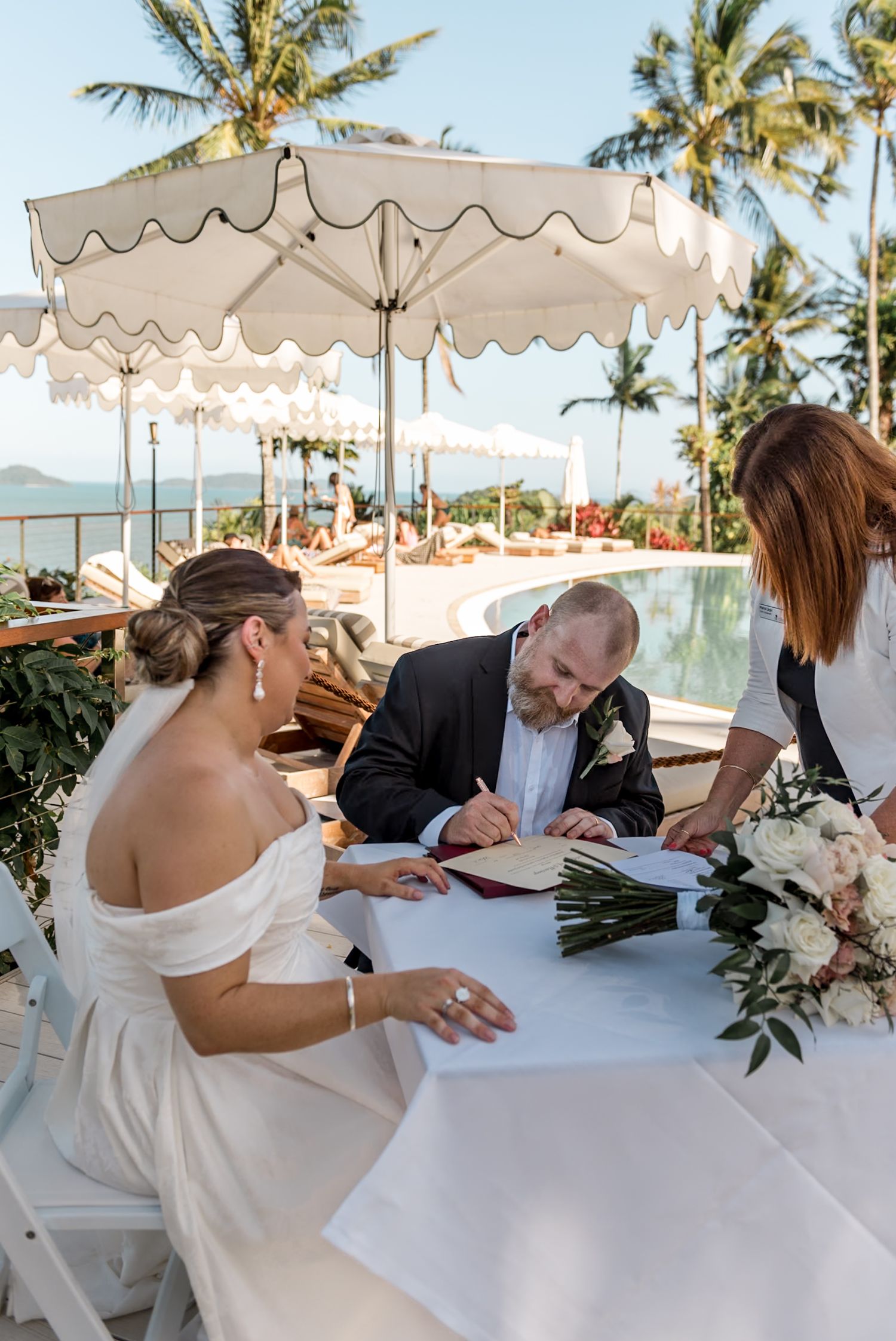 Bride and groom signing marriage certificate at outdoor table with ocean and palm tree views.