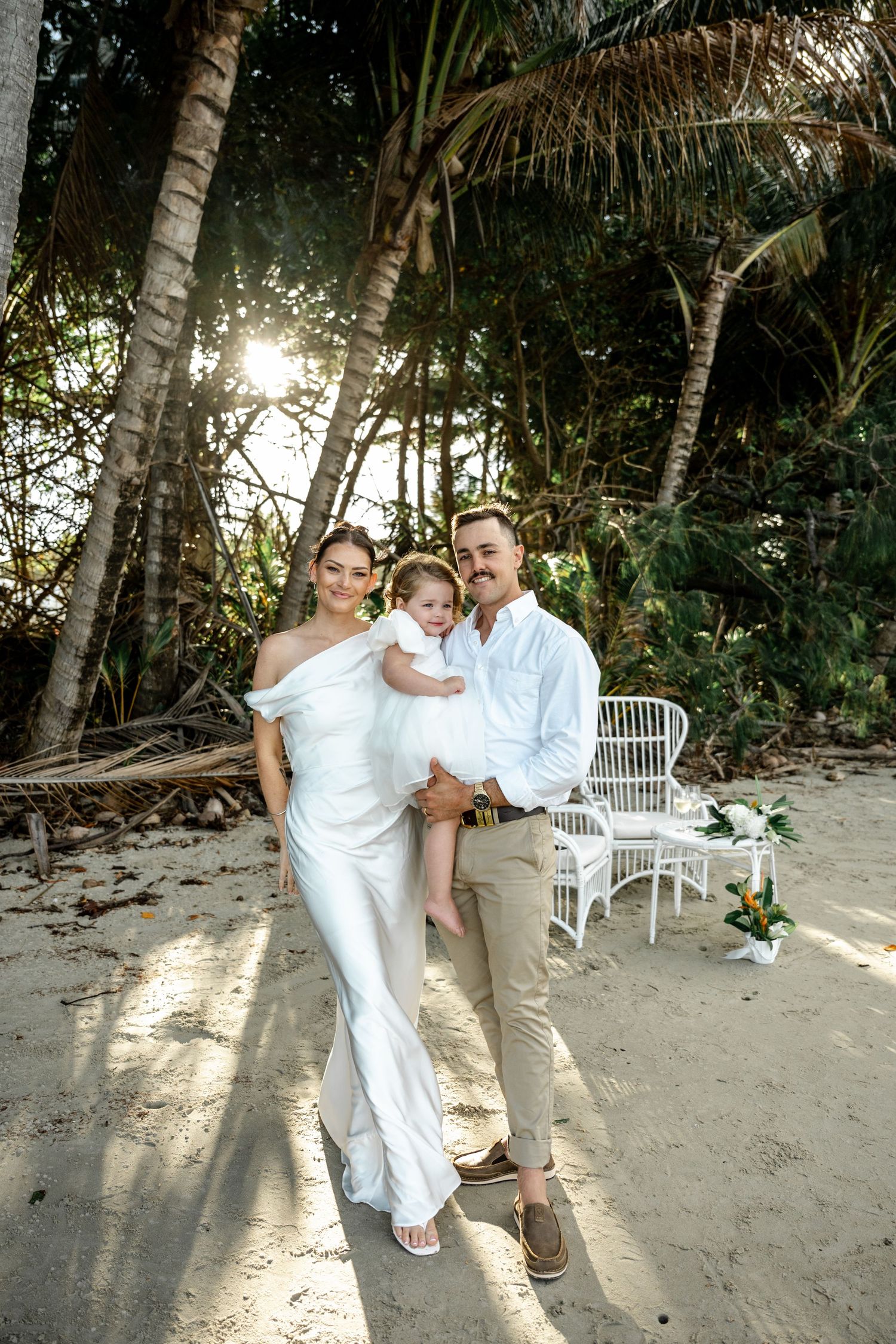 Bride and groom holding their young daughter on a sandy beach surrounded by palm trees at Four Mile Beach, Port Douglas.