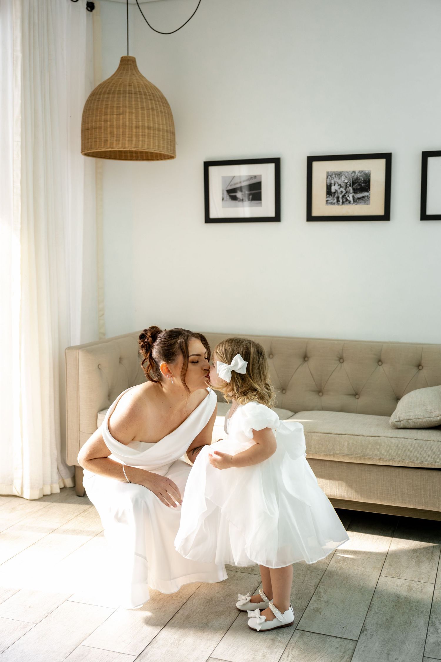 Bride in white wedding gown crouching to kiss a young flower girl in white dress in bright modern room at Sheraton Grand Mirage Resort.