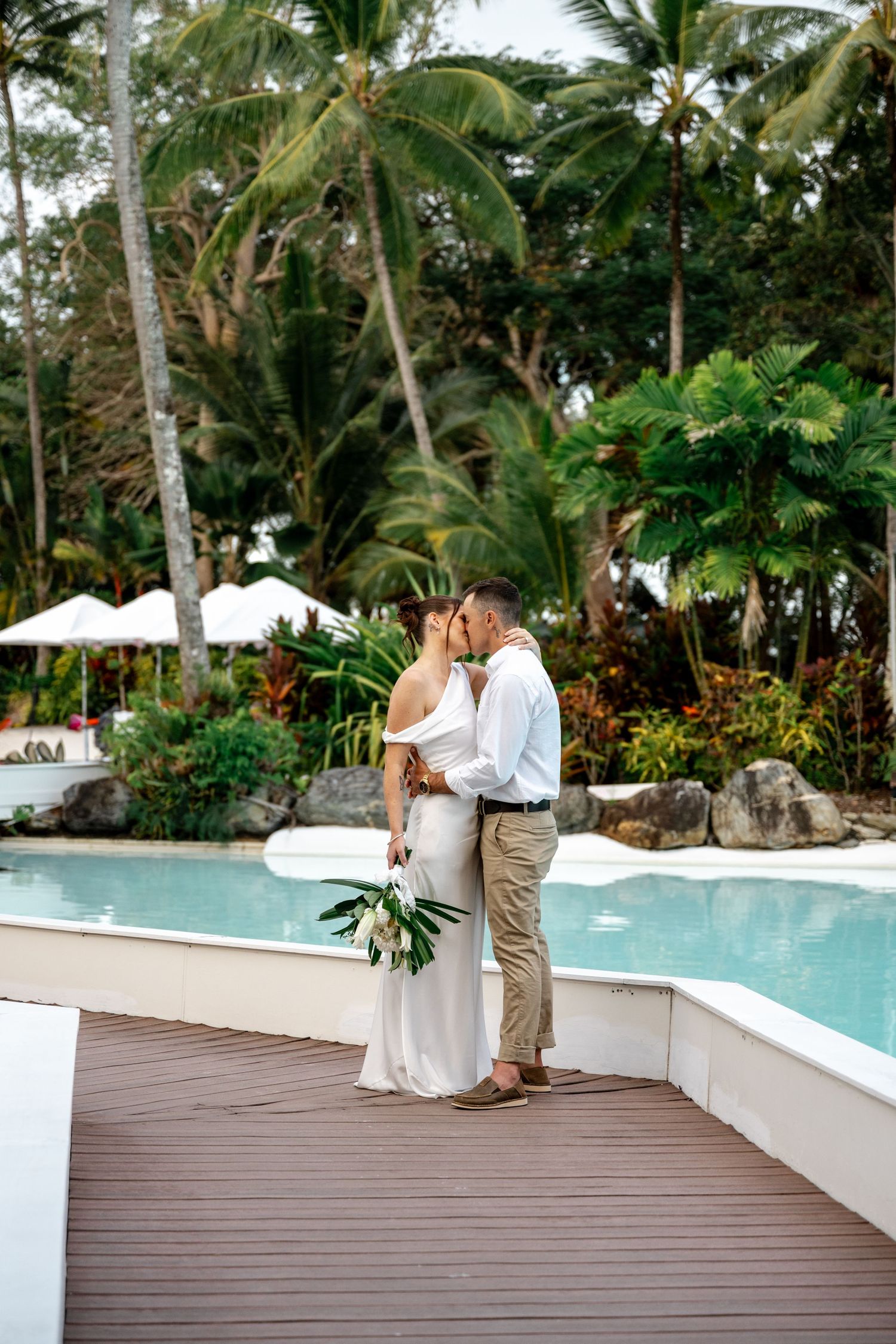 Newlyweds sharing a kiss on a wooden deck beside a tropical resort pool surrounded by palm trees at Sheraton Grand Mirage Port Douglas.