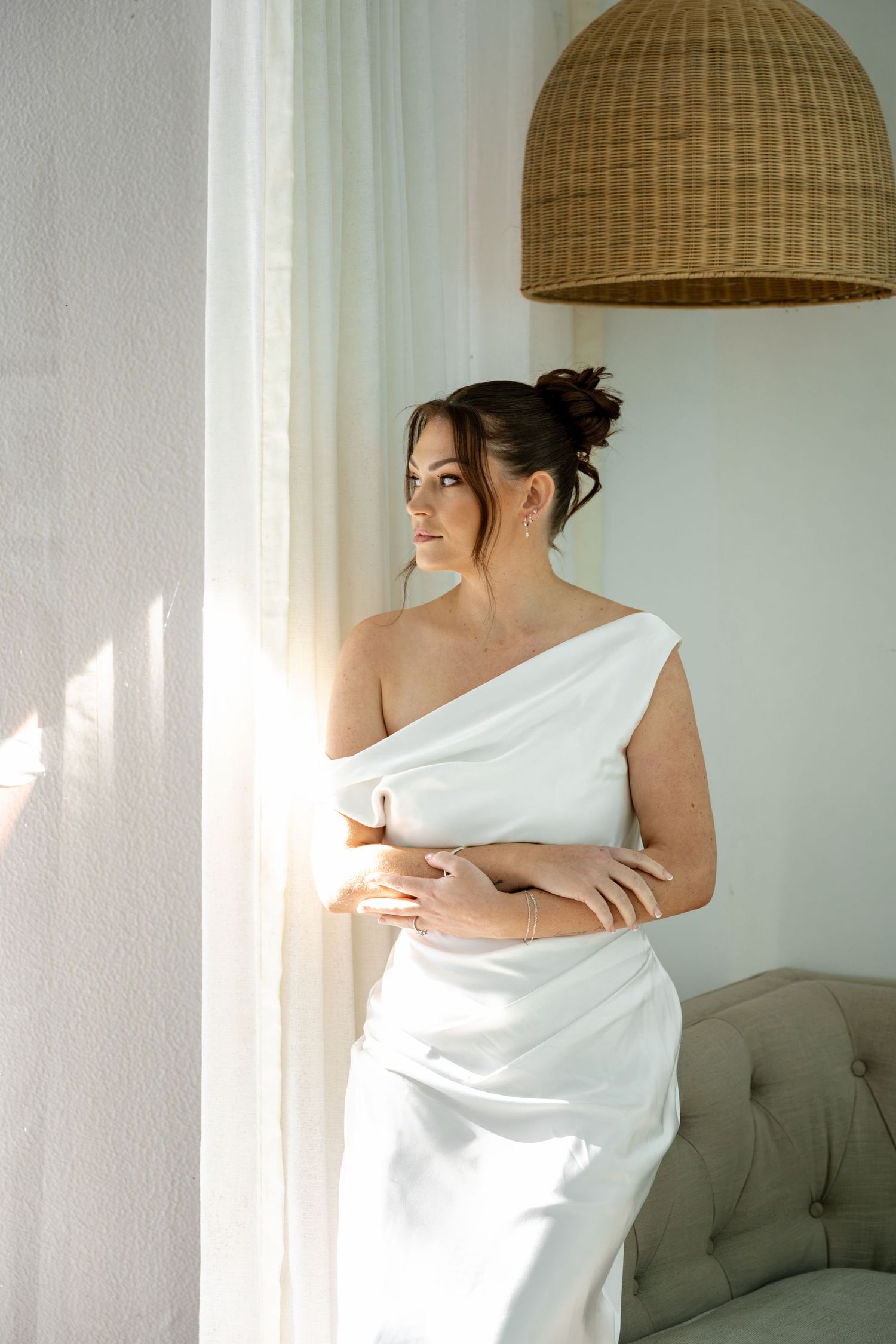 Bride in white one-shoulder satin gown stands by curtained window with arms crossed in sunlit bright room.