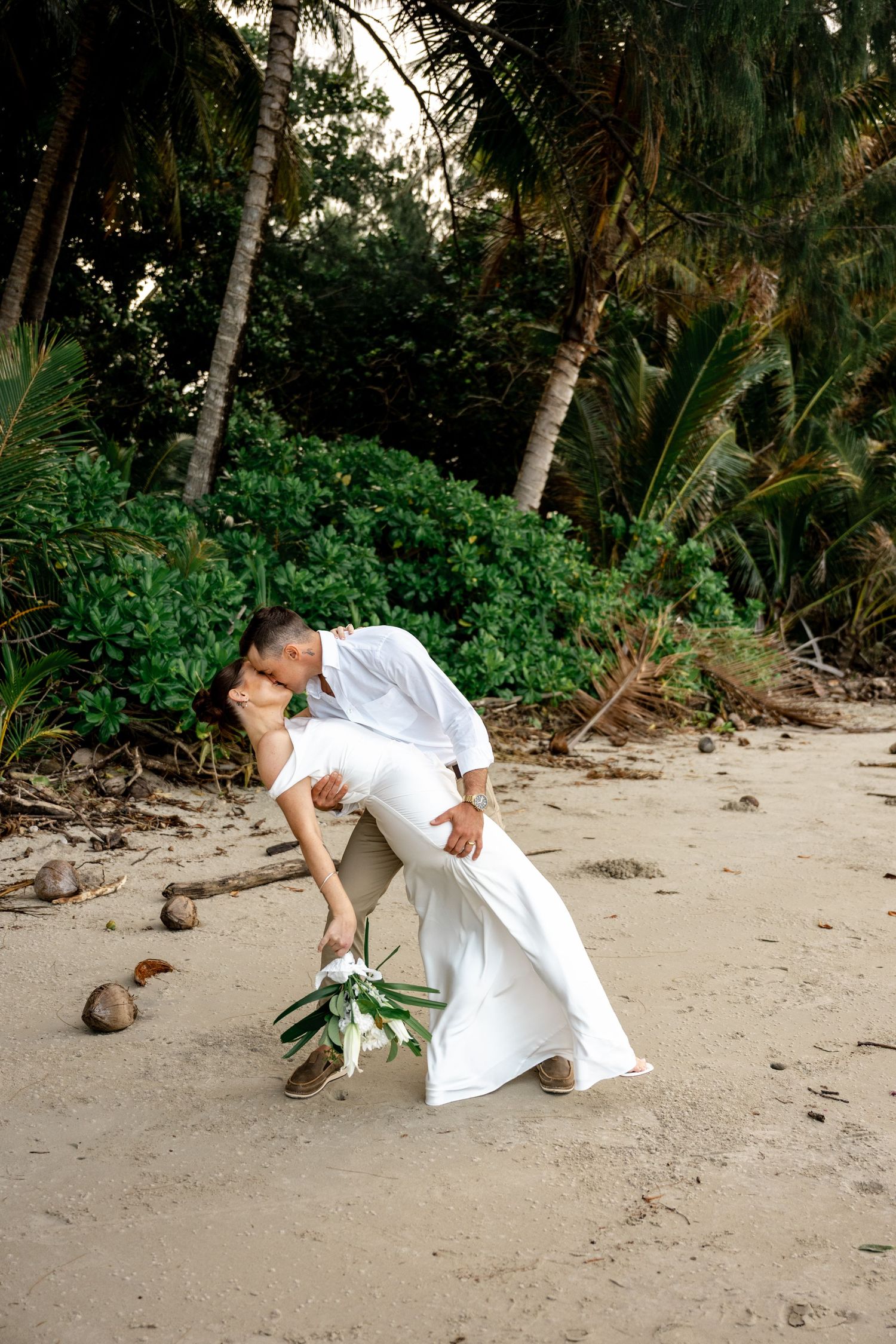 Groom dips bride for a romantic kiss on sandy tropical beach with lush palm trees at Four Mile Beach, Port Douglas.