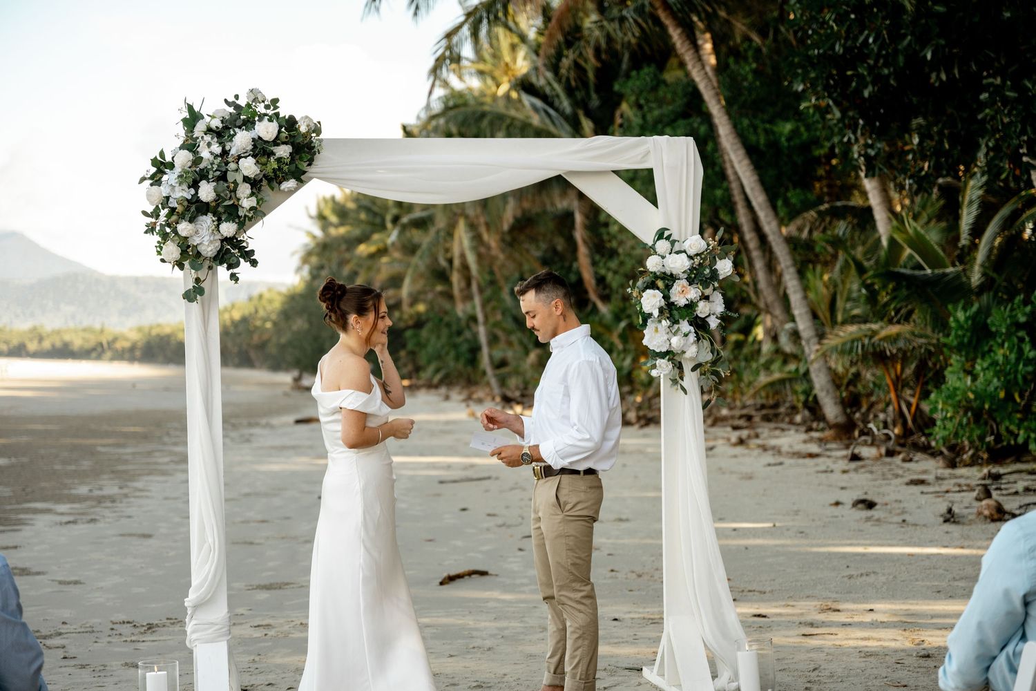 Couple exchanging vows at a beach wedding ceremony under a white floral arch with palm trees in the background at Four Mile Beach.