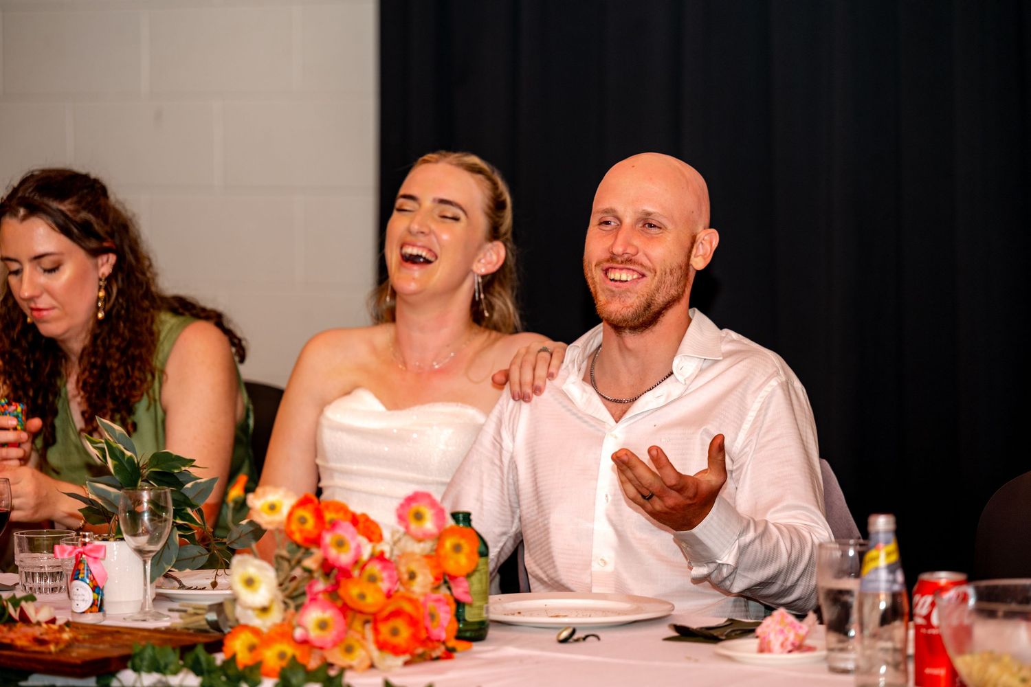 Bride and groom laughing together at wedding reception table adorned with bright orange and pink floral centerpieces.