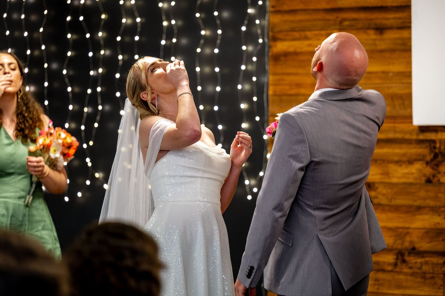 Emotional bride in white veil wiping tear during wedding ceremony with fairy light backdrop.