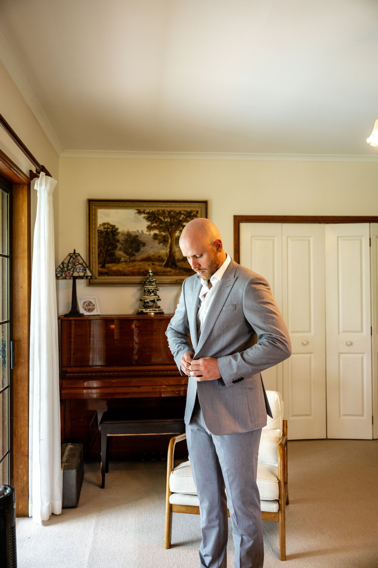 Dapper bald groom buttoning grey suit jacket while getting dressed for wedding ceremony.