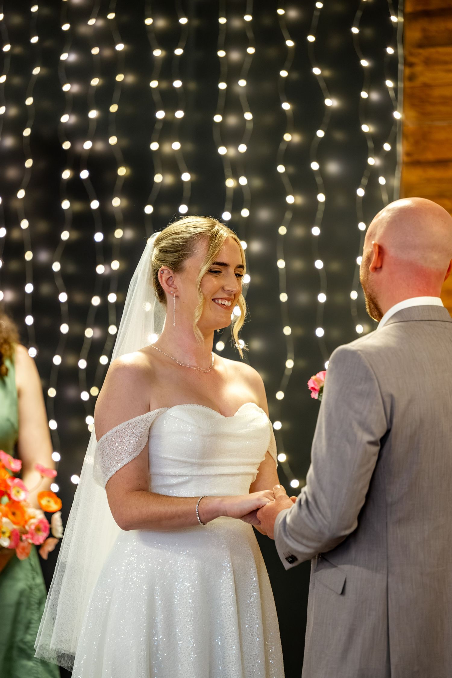 Bride in white gown and veil smiles warmly at groom holding hands before fairy light backdrop at wedding.
