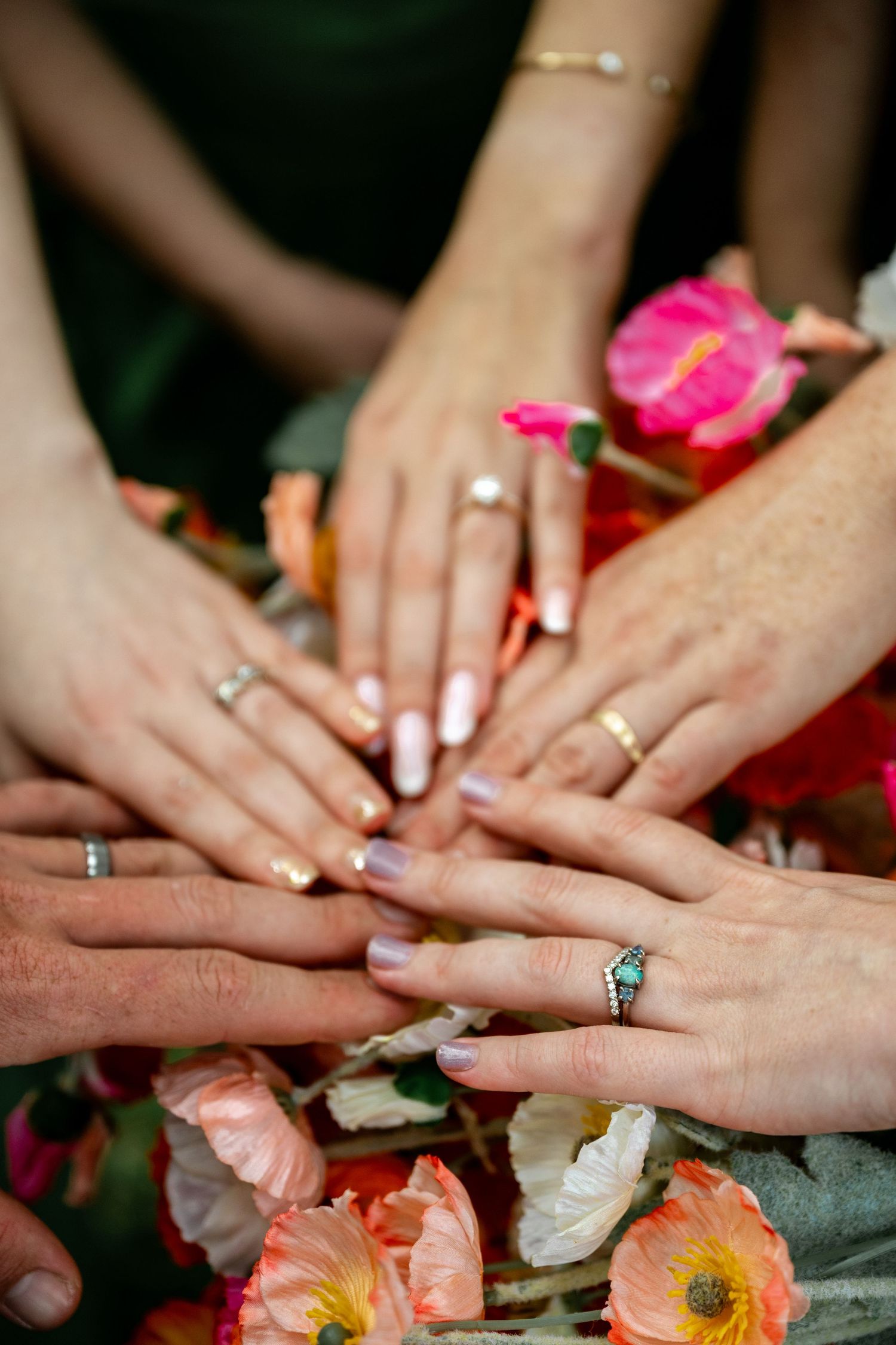 Close-up of wedding party hands displaying rings over vibrant pink and orange floral bouquet arrangement.