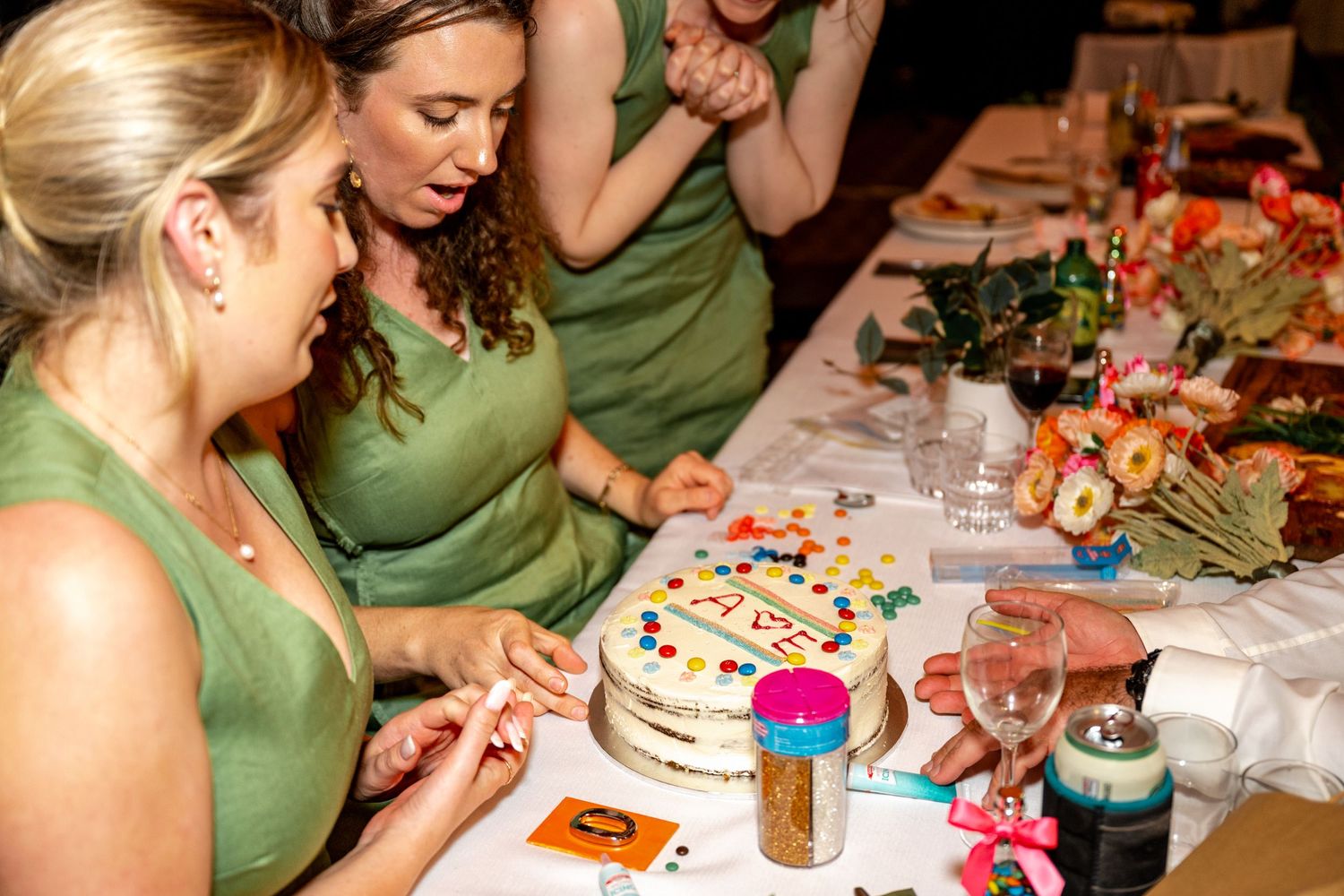 Bridesmaids in green dresses gathered around decorated white cake at wedding reception dinner table.