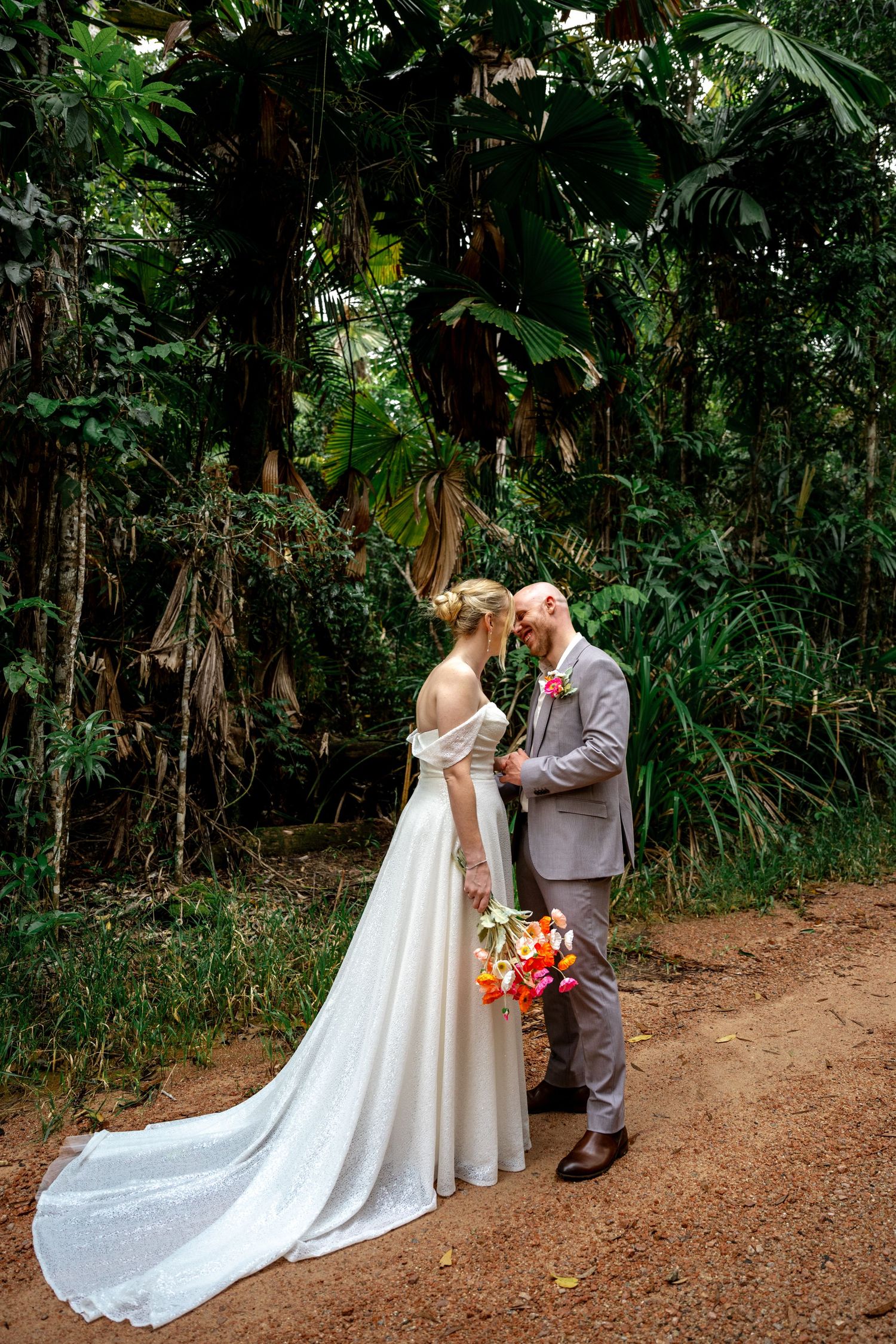 Bride and groom stand forehead-to-forehead on a tropical dirt path, bride showcasing elegant gown train.