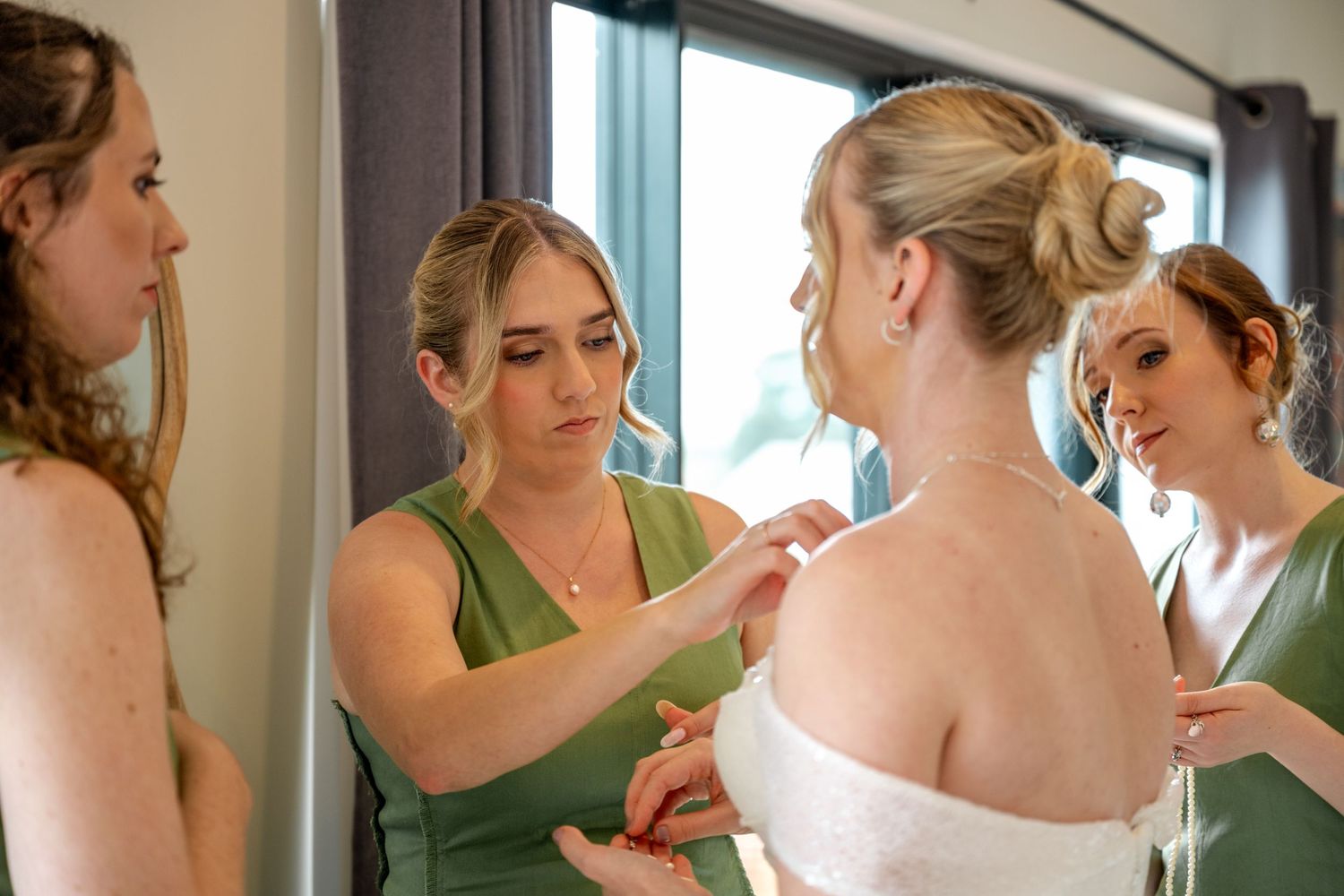 Bridesmaids in sage green help bride with dress details during wedding morning preparations.