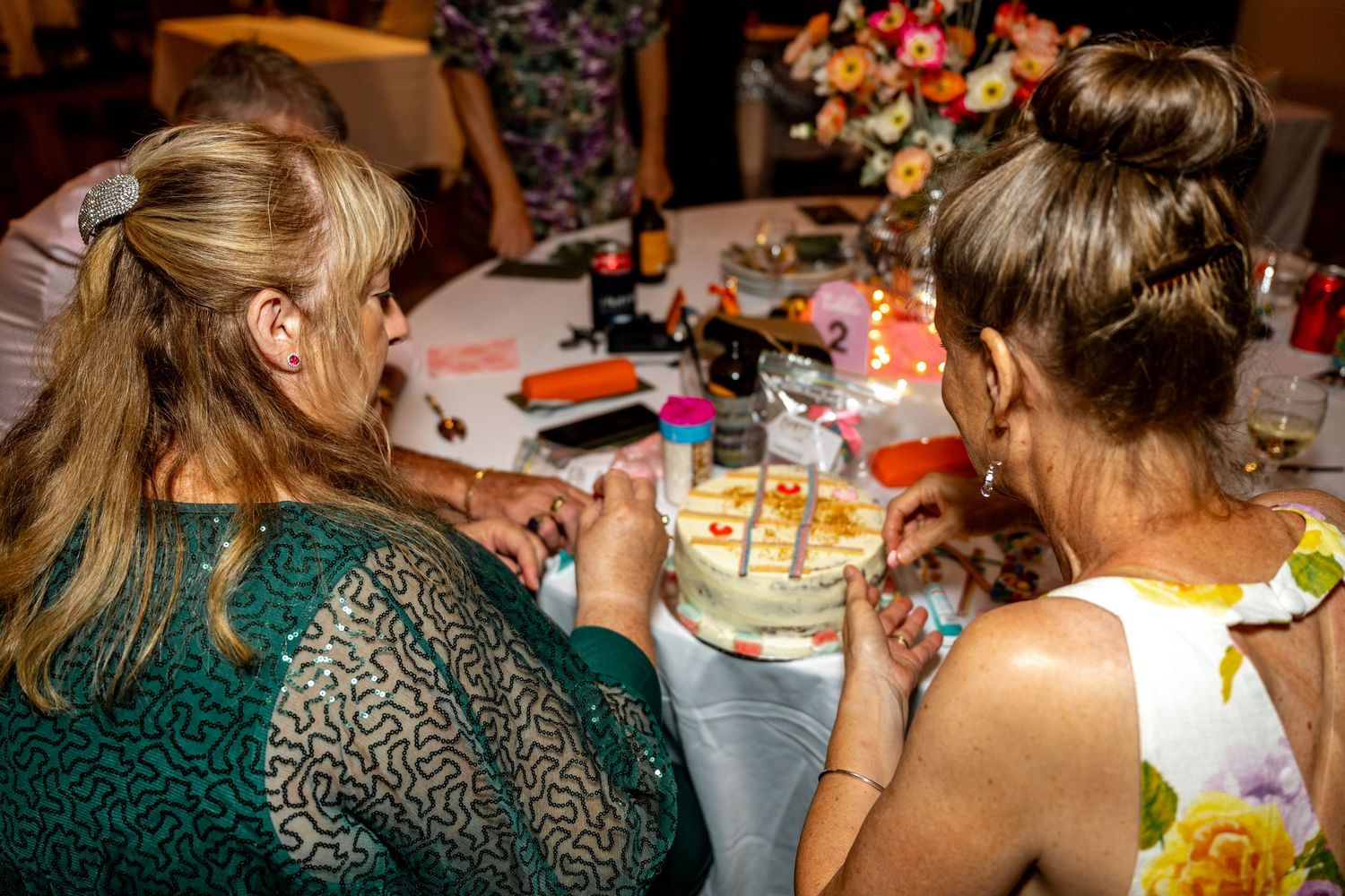 Two women in formal attire decorating a white wedding cake together at a reception dinner table.