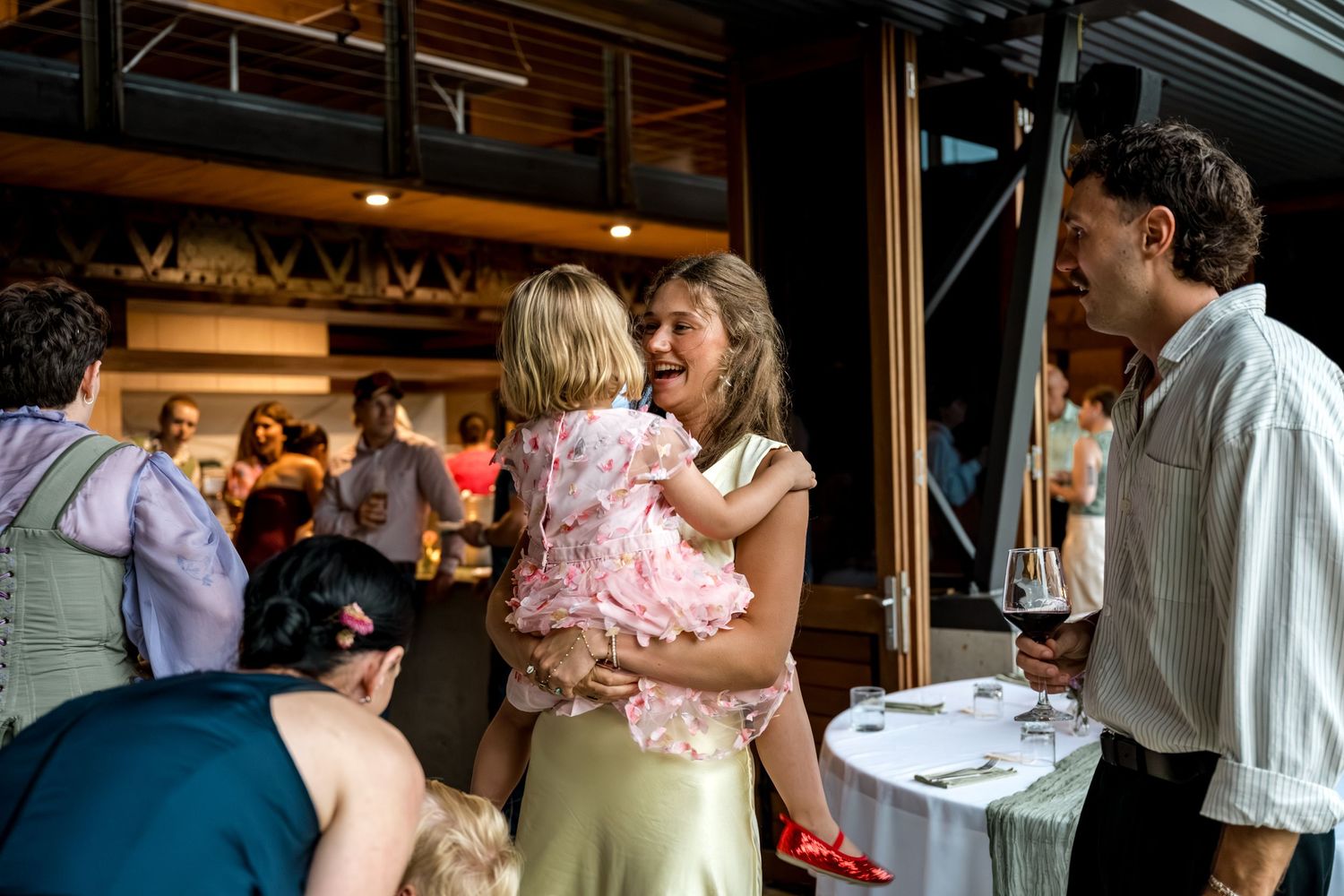 Woman in gold dress joyfully holds young girl in pink at lively wedding reception.