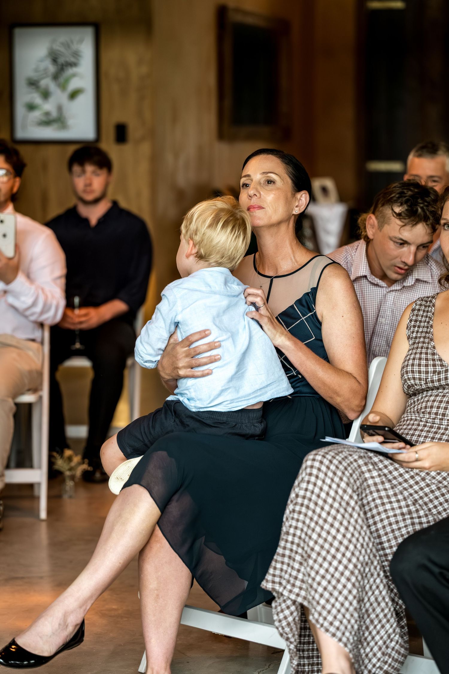 Woman in black dress holding young child on lap while seated at wedding ceremony among guests.