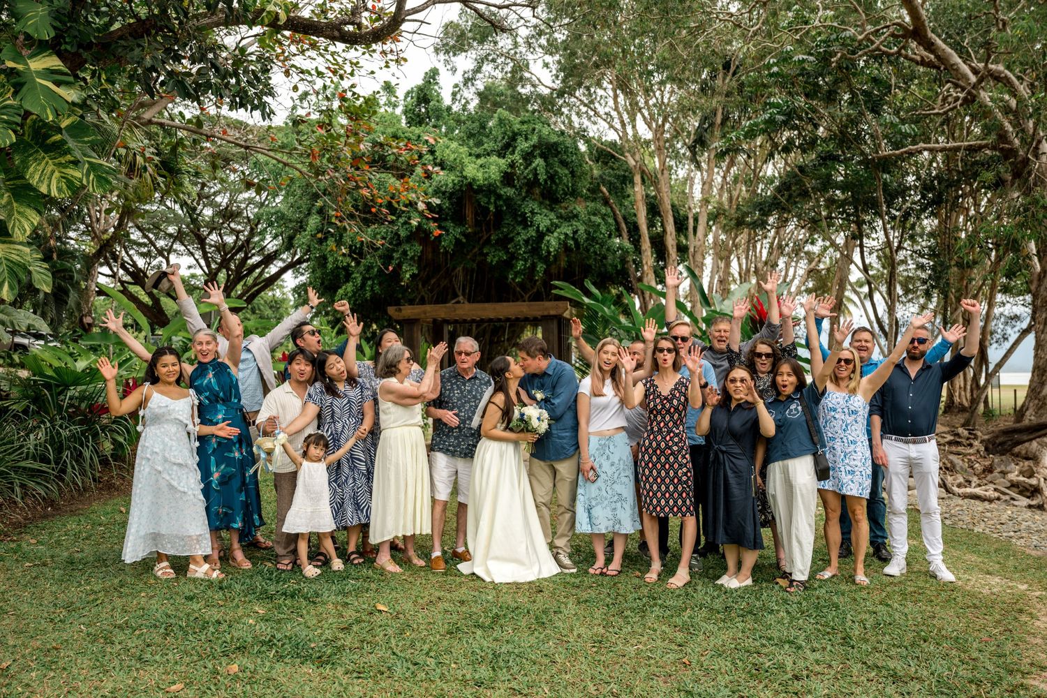 Joyful wedding group photo with all guests cheering and raising arms in tropical garden at Little Cove, Port Douglas.