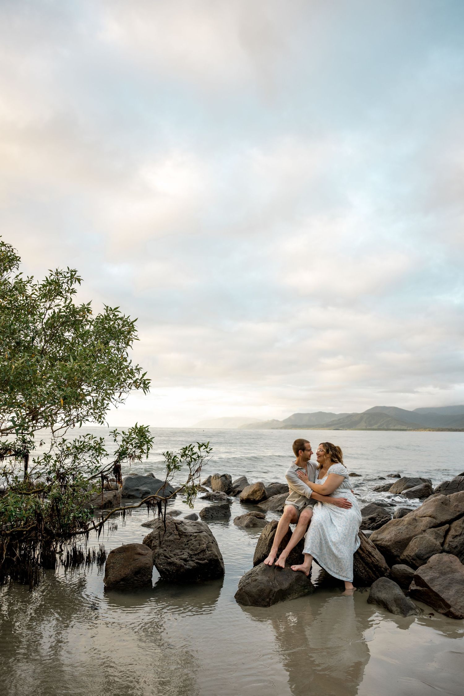 Romantic couple embraces on coastal rocks with calm ocean, mountains, and dramatic cloudy sky behind at Four Mile Beach, Port Douglas.