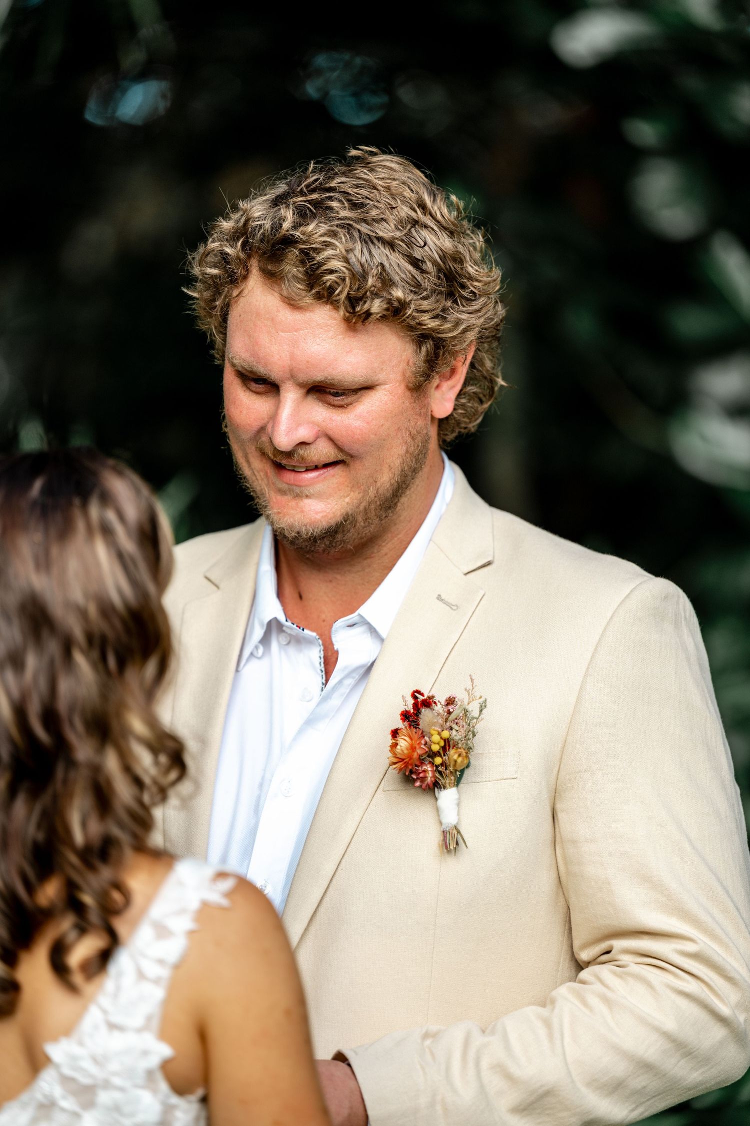 Groom in beige suit smiles warmly during outdoor wedding ceremony with tropical foliage backdrop.