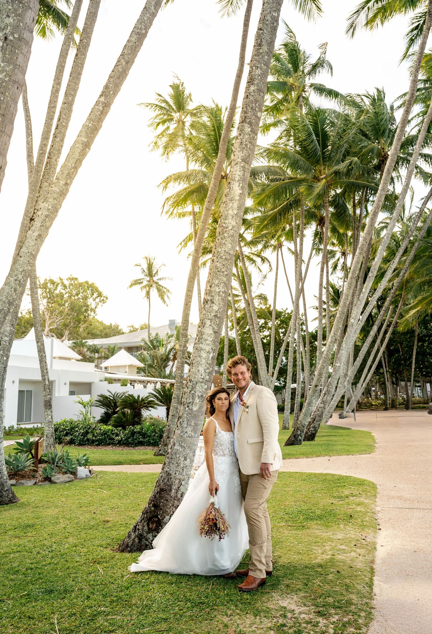 Smiling bride and groom posing together among tall palm trees at a tropical resort on their wedding day.