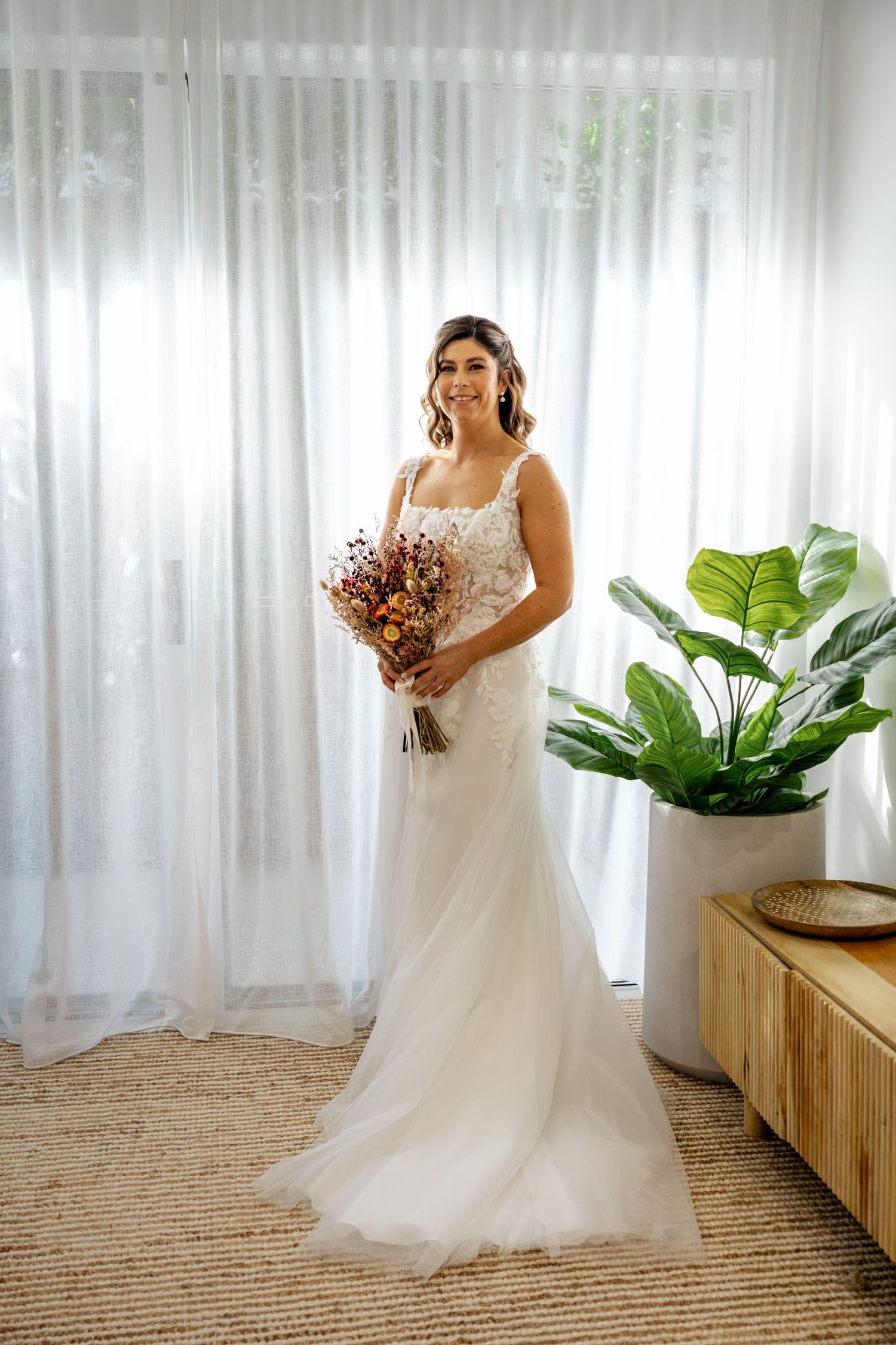 Smiling bride in lace wedding gown holding colorful dried flower bouquet standing by sheer white curtains.