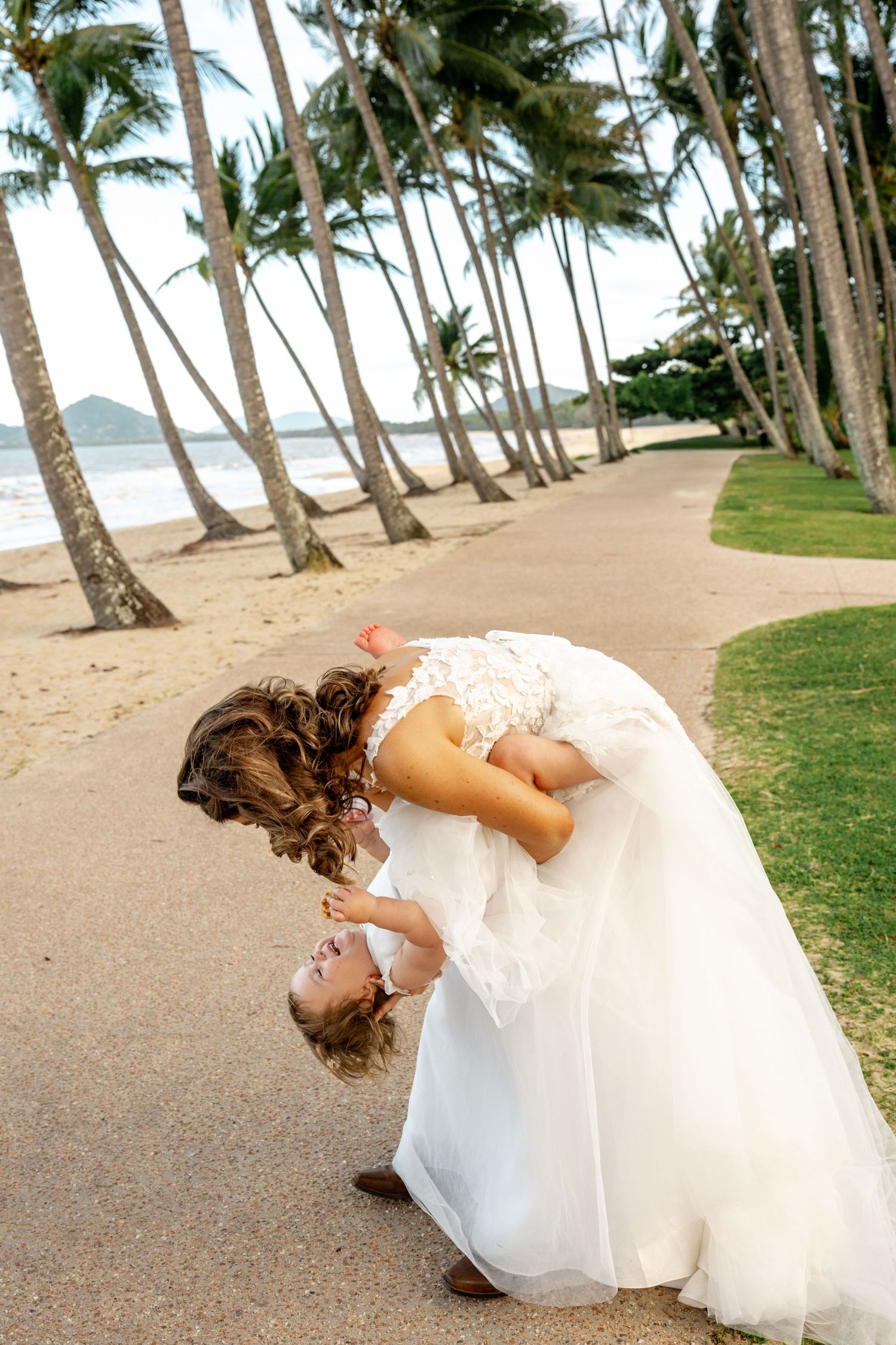 Bride playfully dipping young child upside down on a palm-lined beach path.