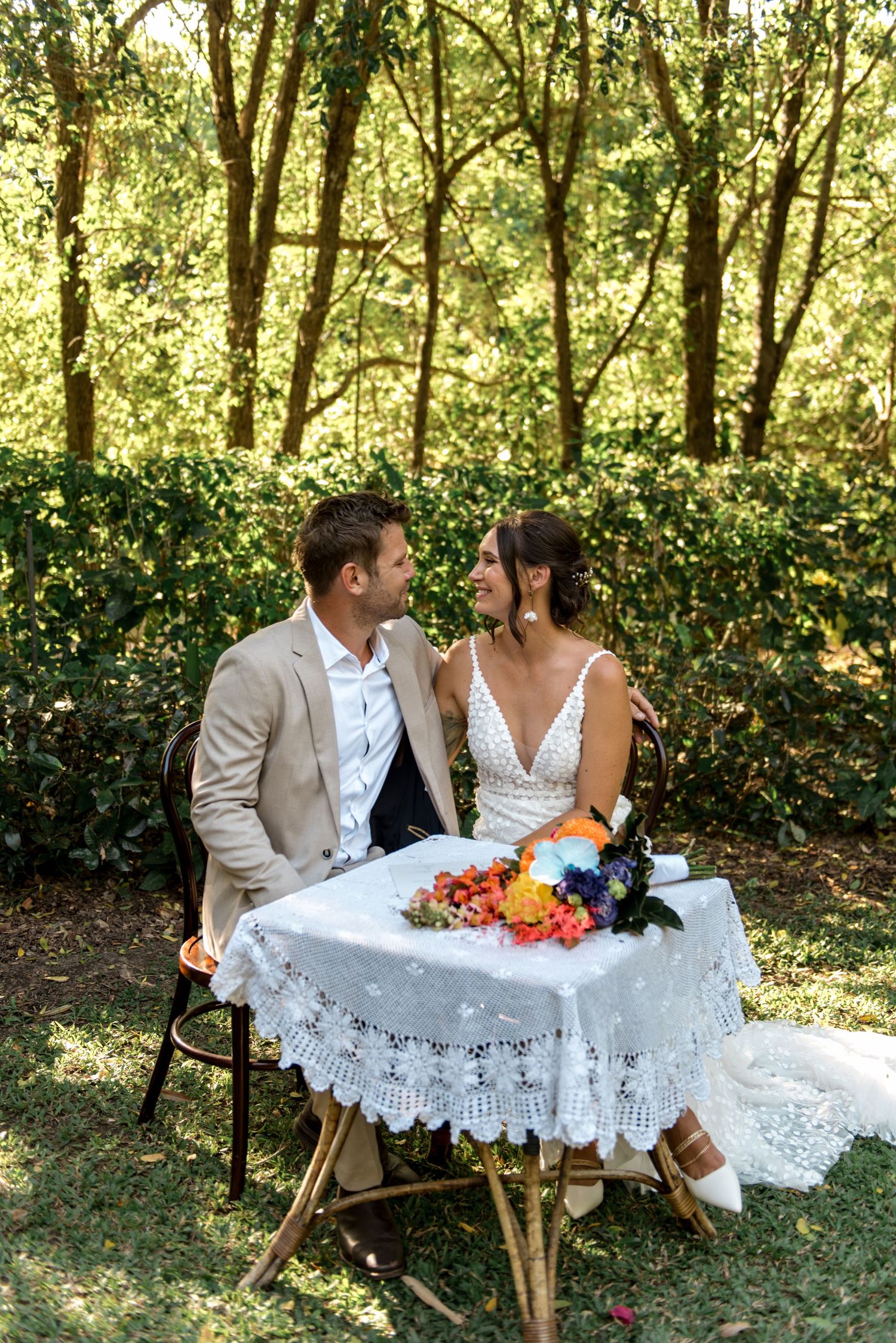 Newlywed couple sits together at lace-covered table in garden, sharing a romantic moment after ceremony.
