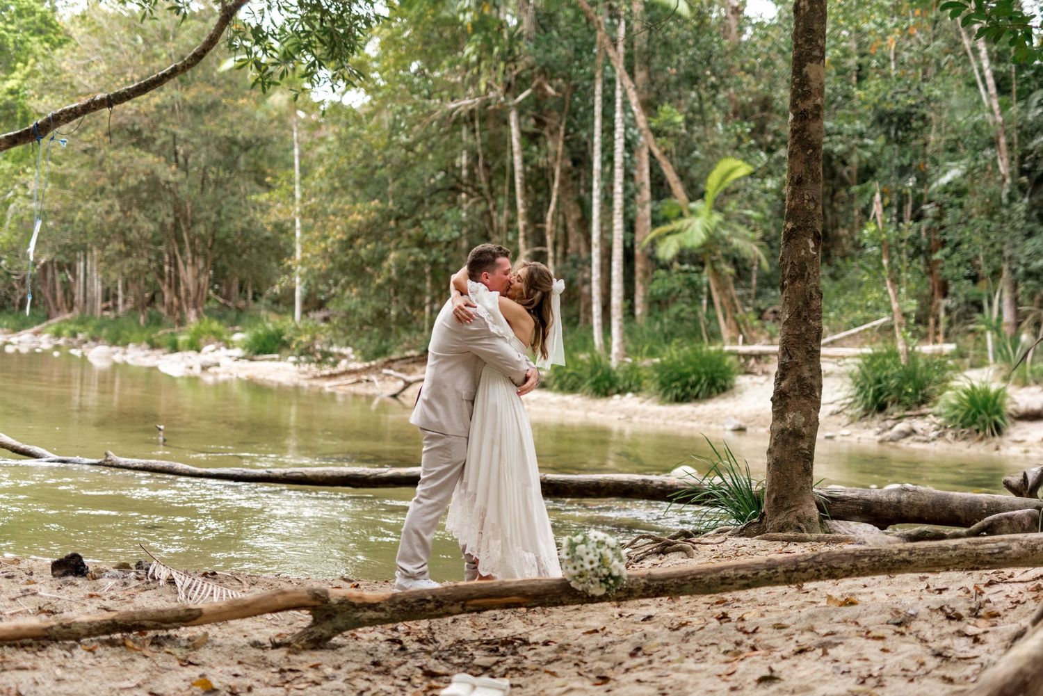 Bride and groom share a romantic kiss by a serene forest creek on their wedding day.