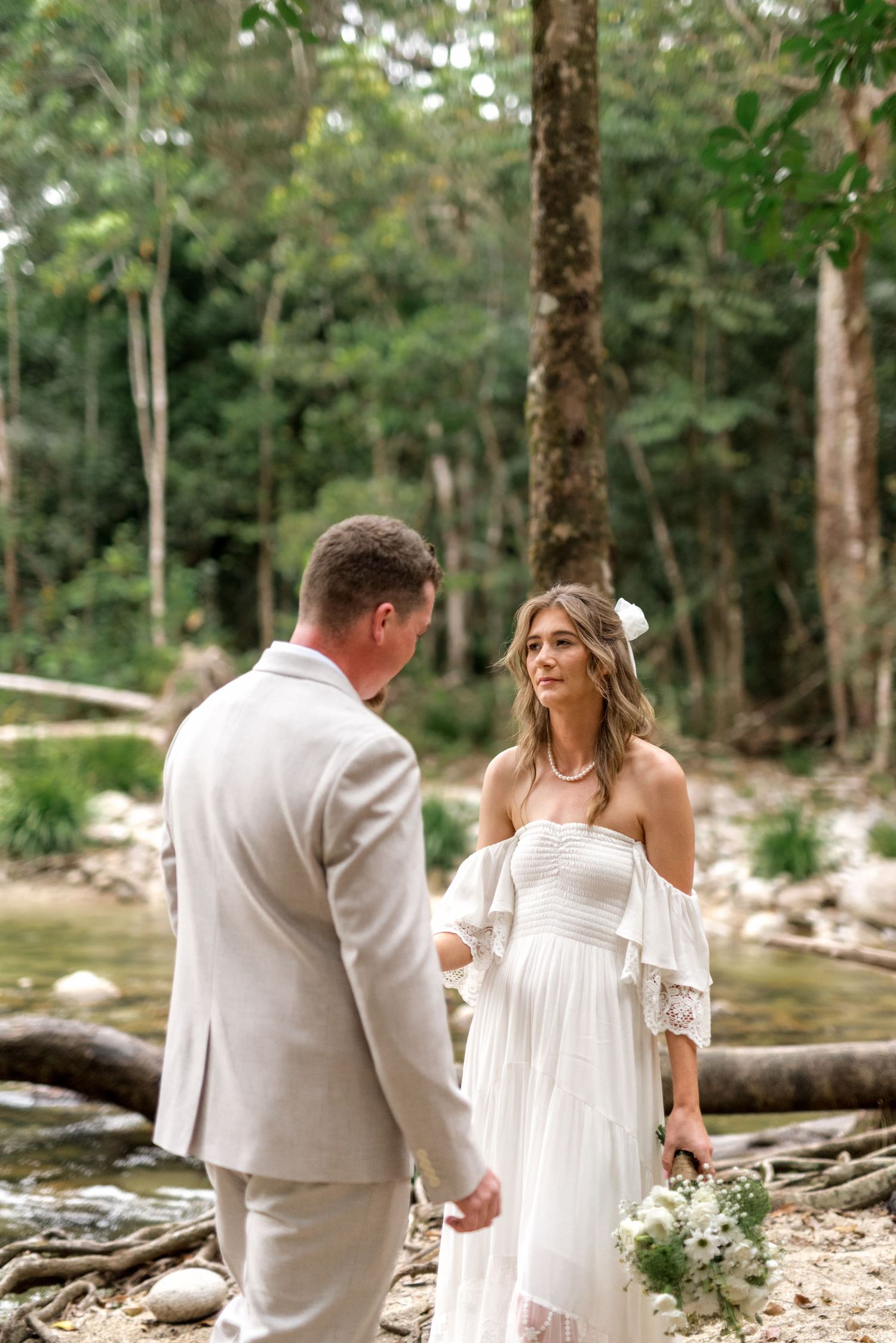 Bride in boho white gown holds bouquet while facing groom by a forest creek.
