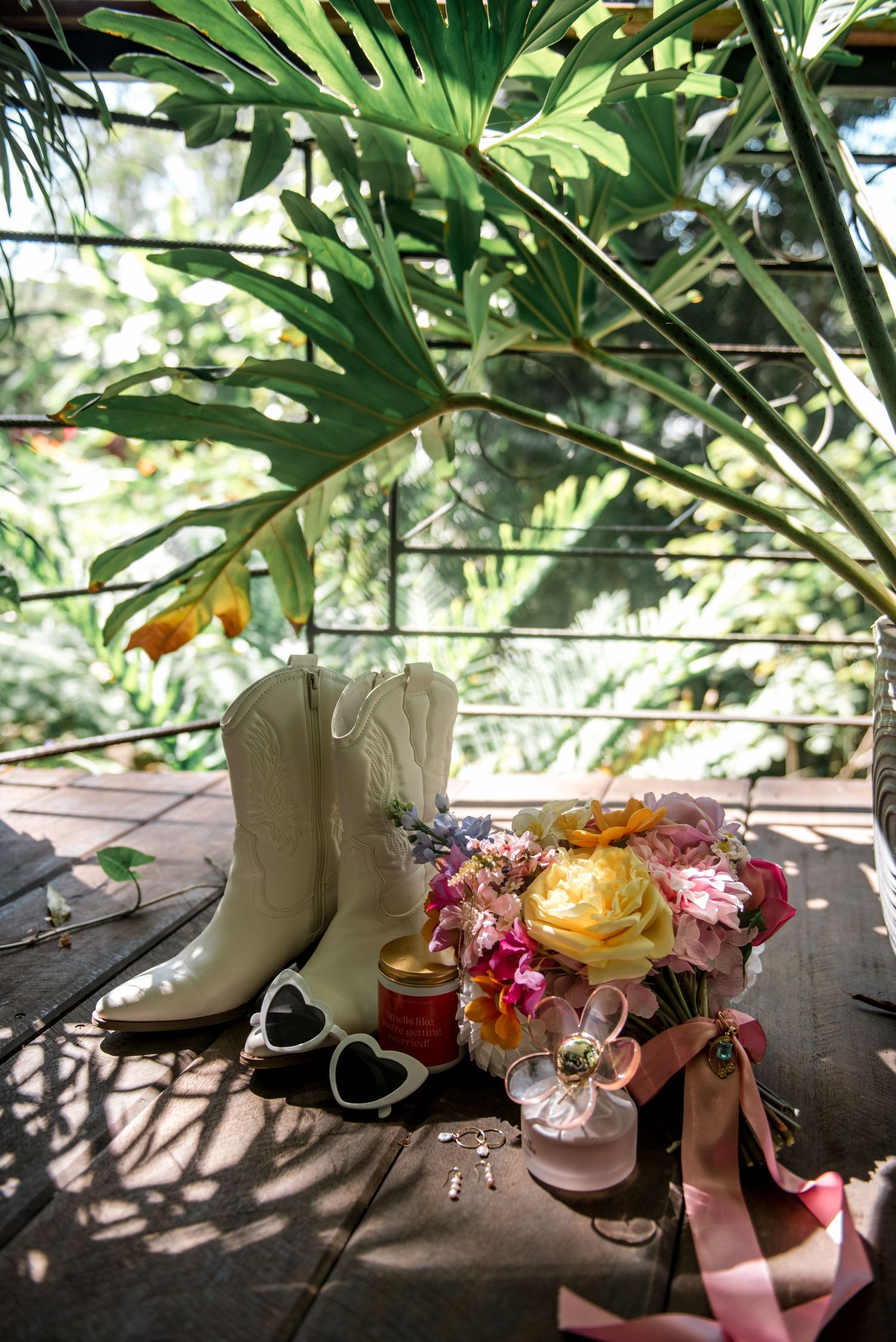 White cowboy boots, colorful bridal bouquet, heart sunglasses, and accessories on wooden surface.