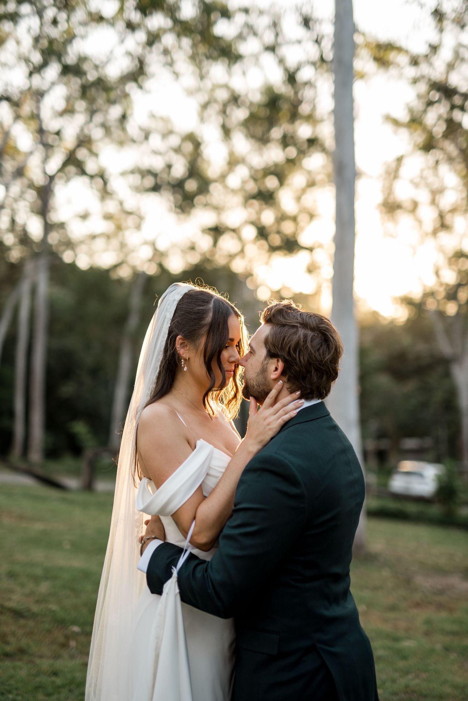Bride in off-shoulder gown and veil embracing groom in dark suit at golden hour.