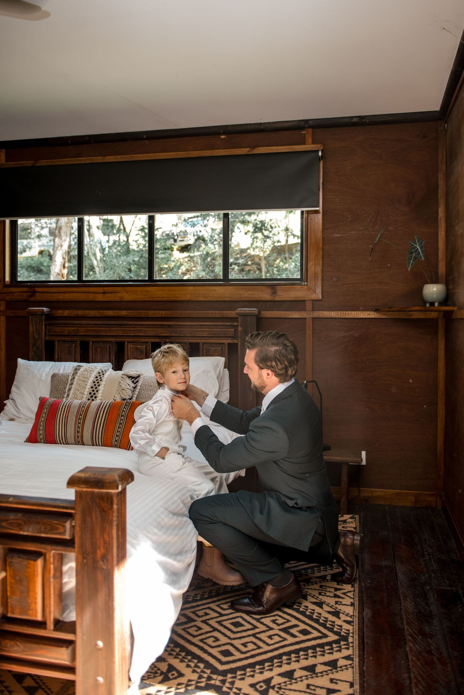 Groom kneeling to adjust young boy's white wedding outfit beside a rustic wooden bed with patterned rug.
