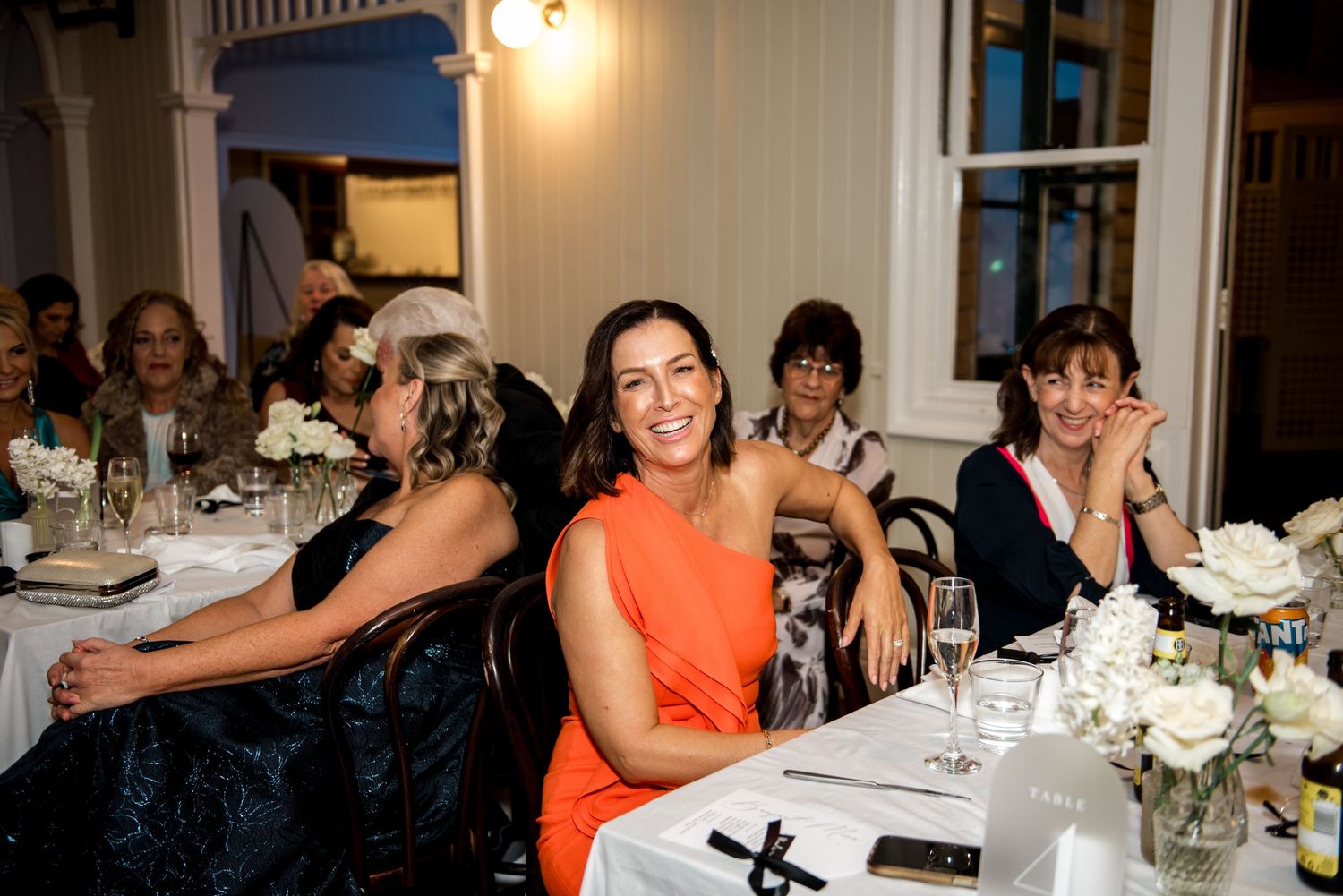 Smiling wedding guests in elegant attire seated at decorated reception tables with white flowers.