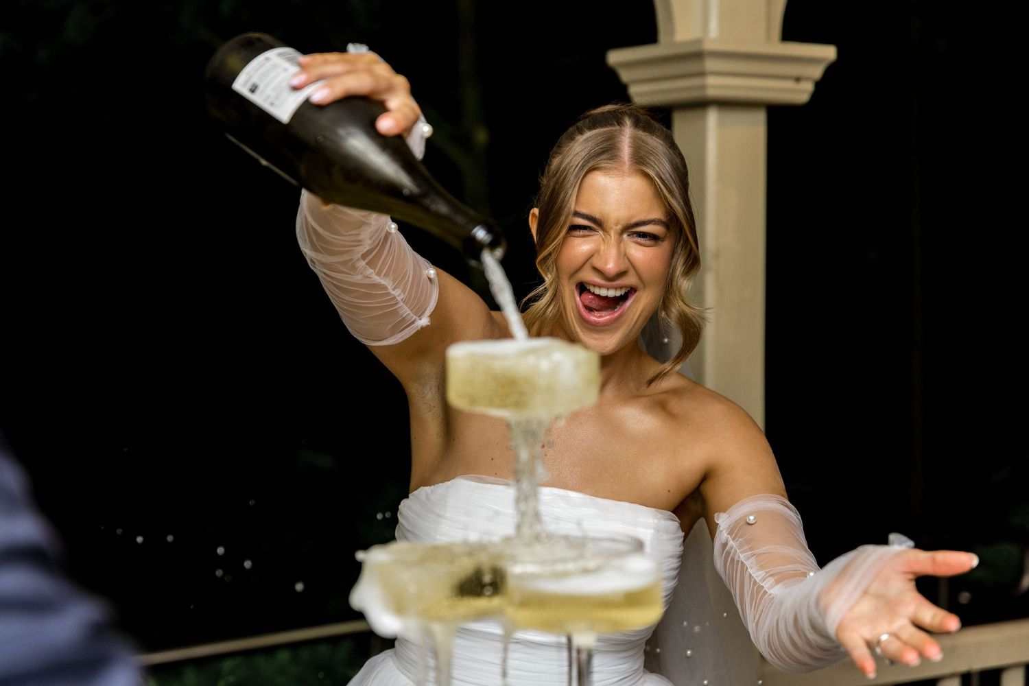 Excited bride laughs as champagne overflows from a glass tower at her wedding.