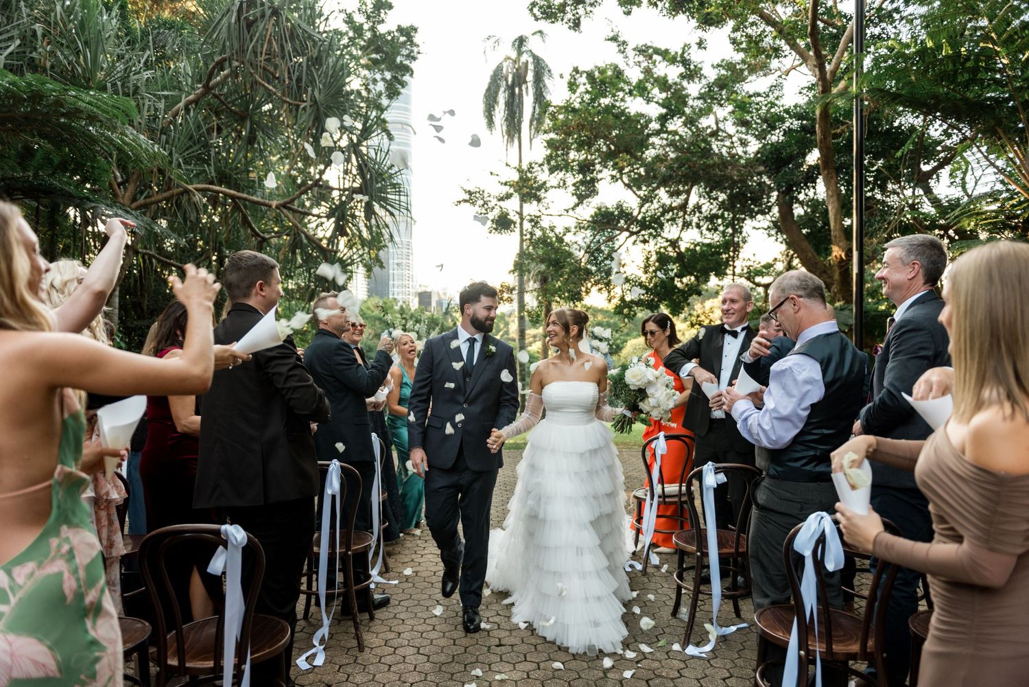 Bride and groom walking down outdoor aisle as guests celebrate with confetti at wedding.