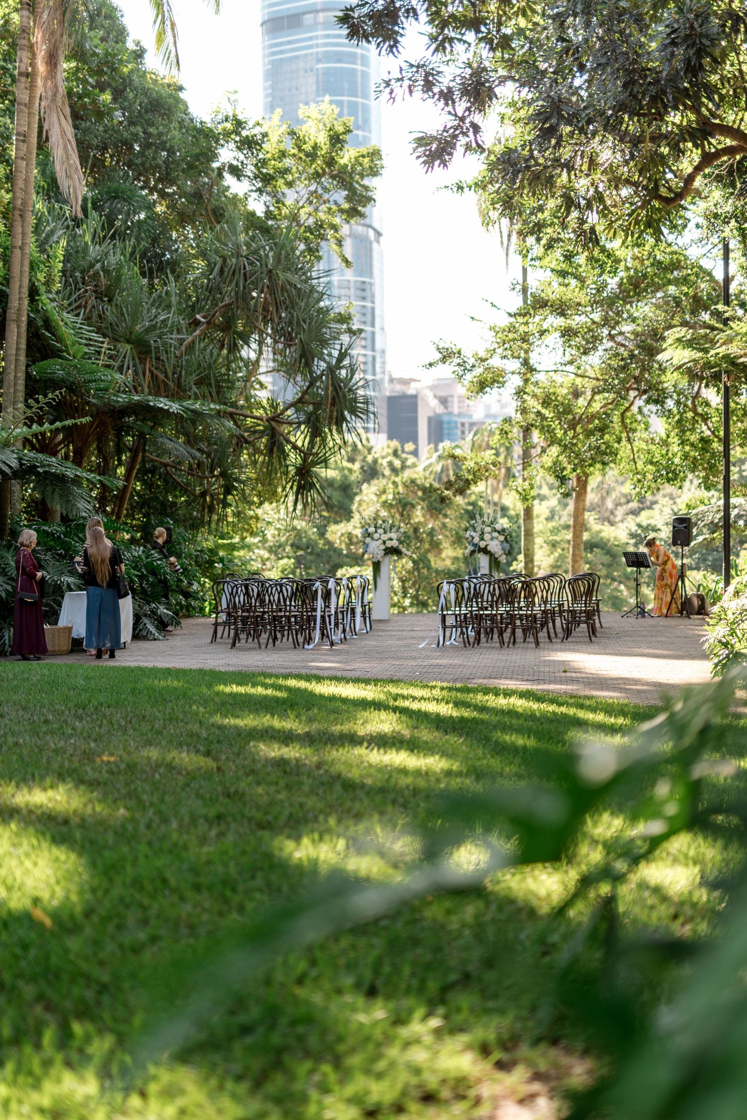 Wide view of outdoor wedding ceremony venue with bentwood chairs set among tropical gardens and city skyline.